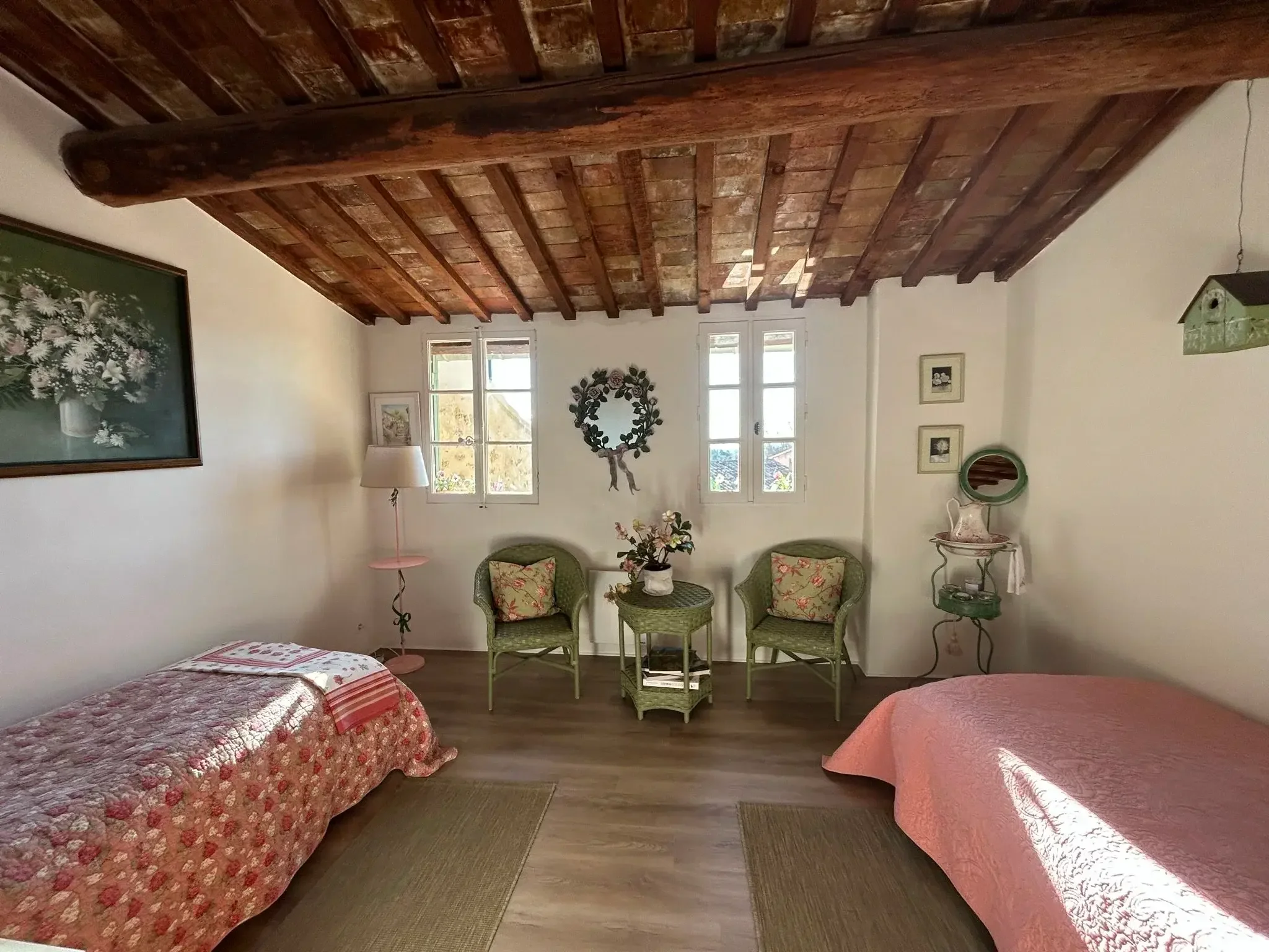 Chambre avec deux lits aux oreillers à motifs floraux, un mur blanc avec des fenêtres, un plafond en bois avec poutres apparentes, une table basse avec des plantes, et des fauteuils verts.
