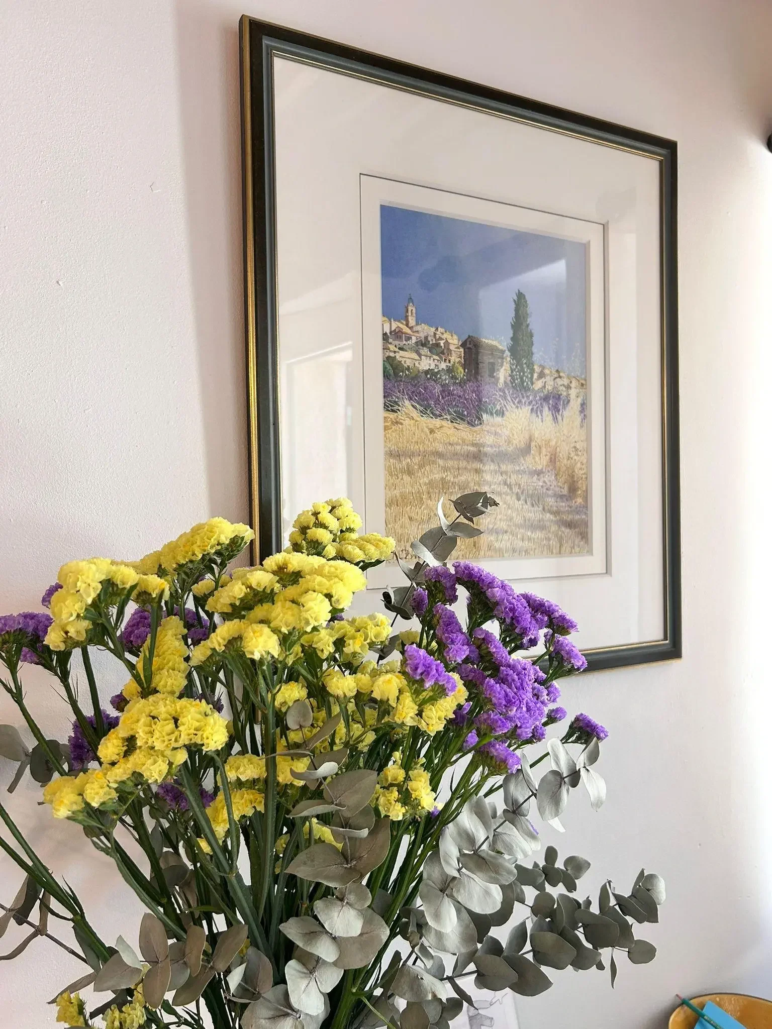 Un bouquet de fleurs jaune et violette placé devant un tableau encadré représentant un paysage avec des maisons, des collines et un ciel bleu, accroché à un mur blanc.