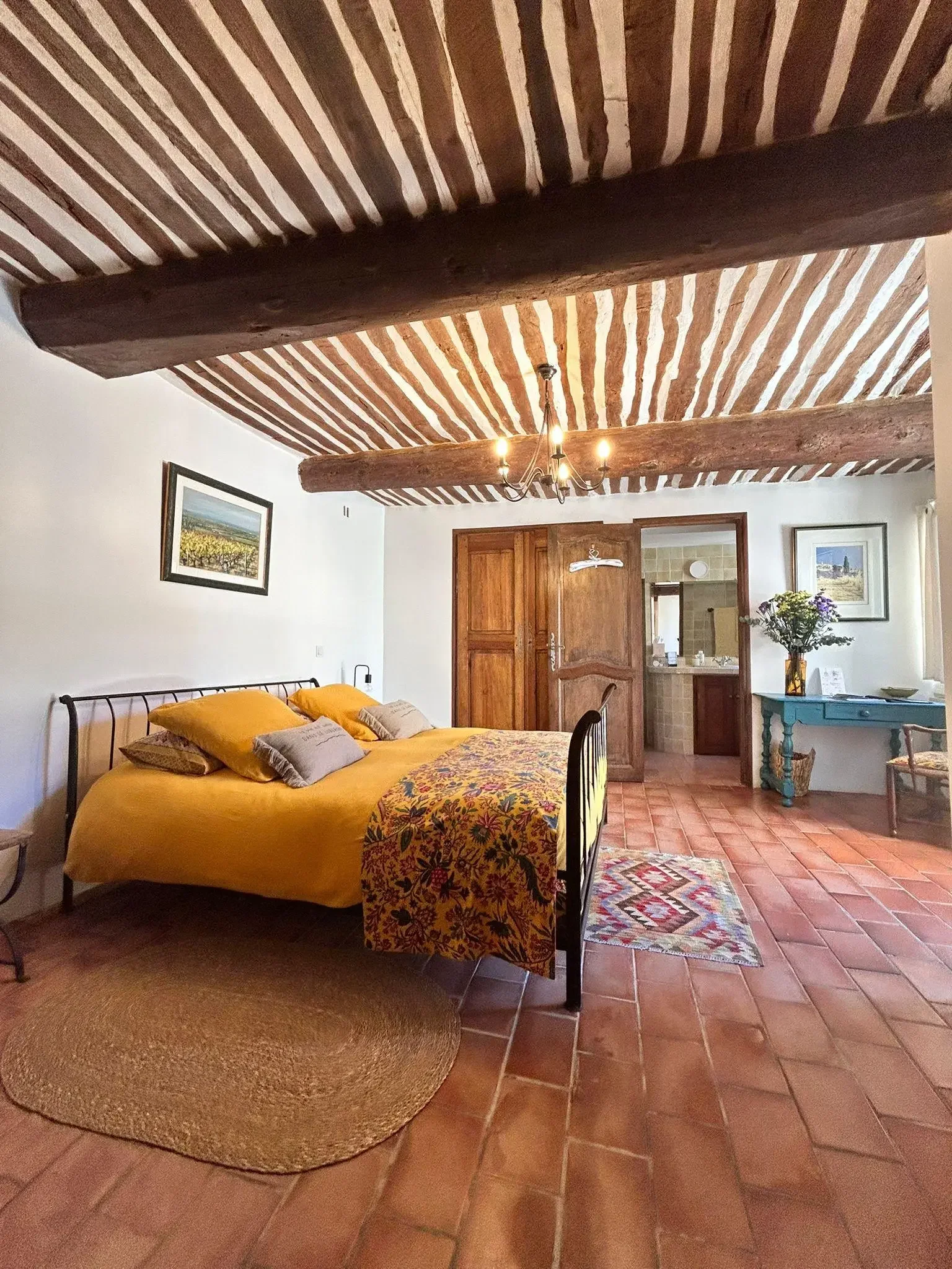 Chambre aménagée avec un lit en fer avec une couverture jaune et plusieurs oreillers, un tapis rond en paille, une commode bleue avec un vase de fleurs, des tableaux sur le mur, un plafond en bois avec poutres apparentes et un lustre, porte en bois m