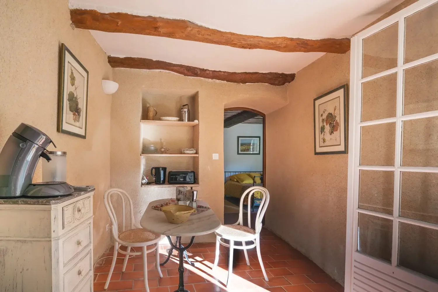 Intérieur d'une cuisine rustique avec un mur en terre cuite, poutres en bois au plafond, un petit table ronde avec deux chaises, et une étagère avec divers appareils électriques.