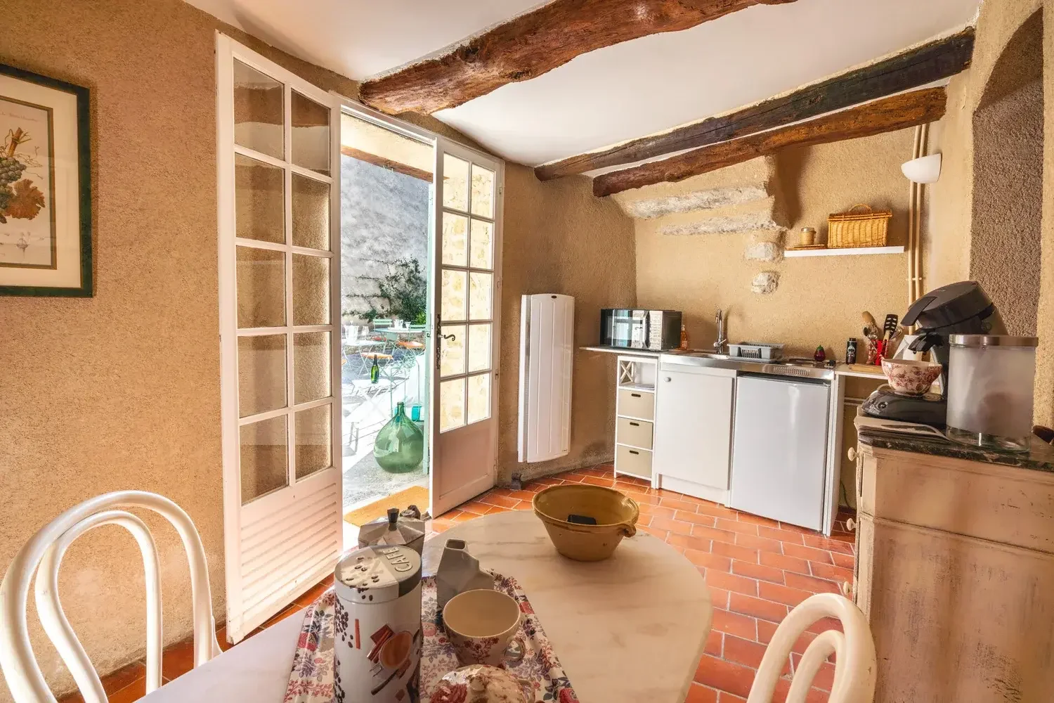 Cuisine rustique avec murs en terre, poutres en bois au plafond, porte-fenêtre donnant sur un balcon avec plantes et table à manger avec vaisselle.