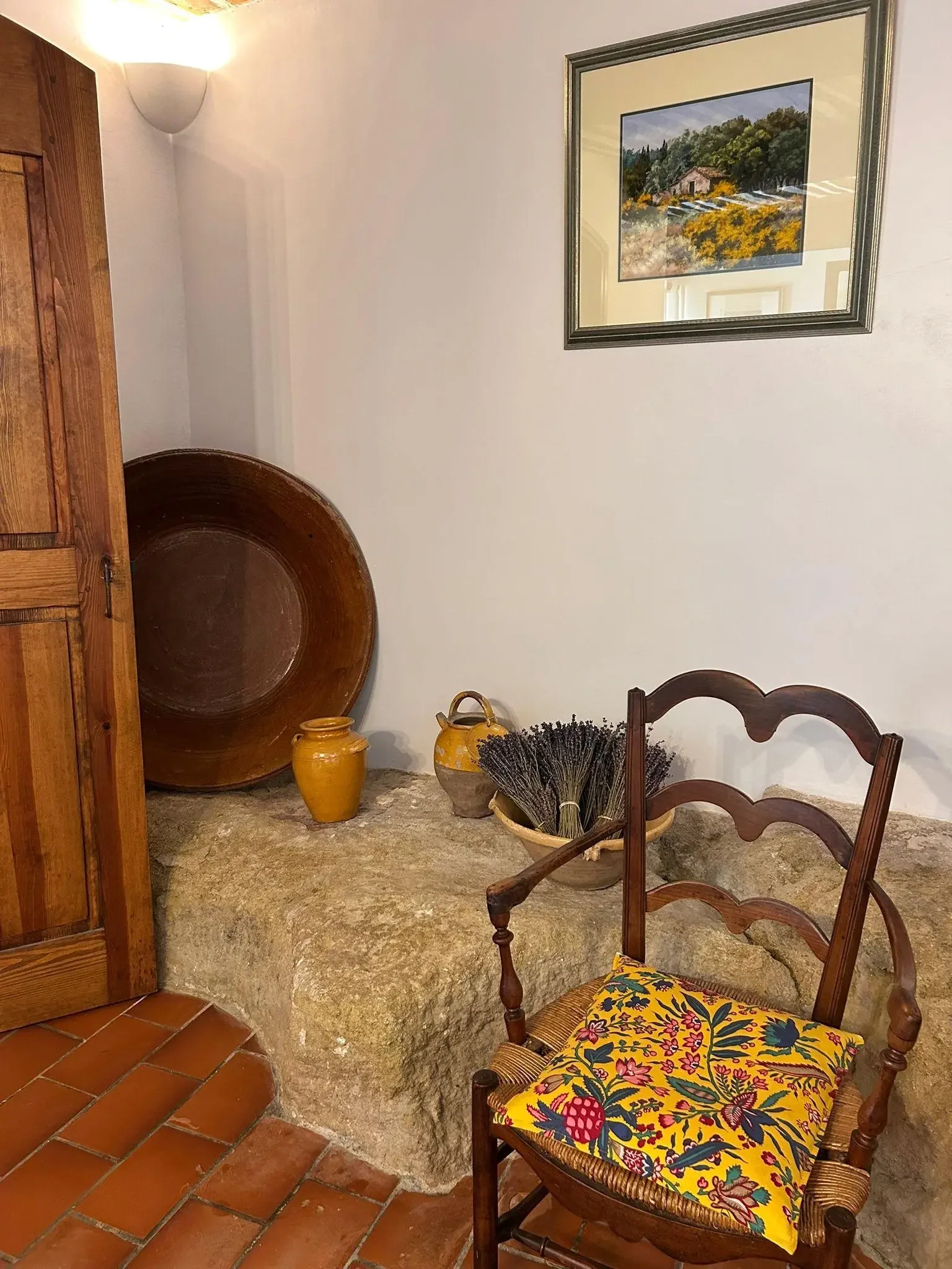 Intérieur rustique avec un fauteuil en bois doté d’un coussin fleuri, un panier de lavande, plusieurs poteries jaunes, un miroir et une peinture de paysage dans le fond.