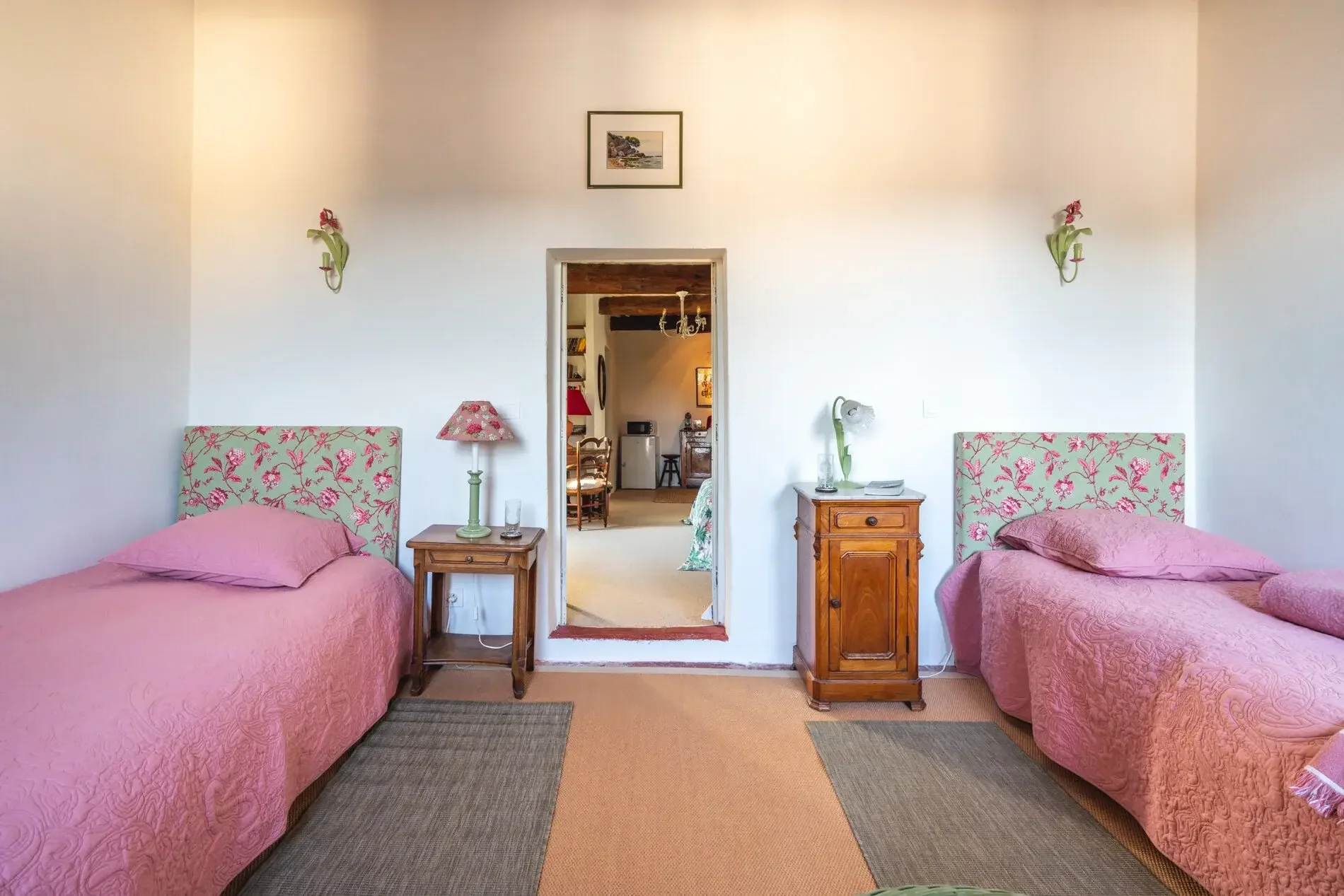 Chambre avec deux lits simples, têtes de lit fleurie, table de nuit avec lampe, dans une maison avec vue sur une chambre et un salon, décoration pastel, mur blanc, tapis au sol.