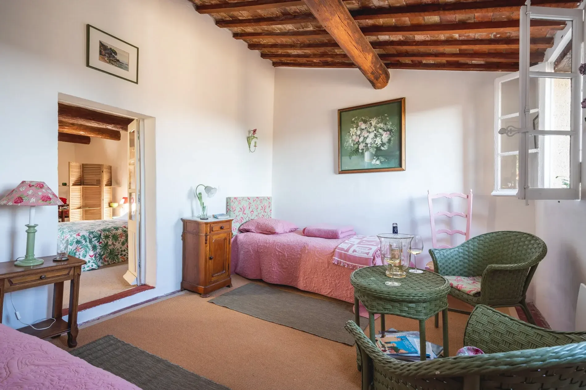 Chambre avec lit simple rose, tableau de fleurs au mur, rideau gris clair, fenêtres ouvertes, meubles en bois, tapis beige, fauteuils en rotin avec verres de vin sur une table, et porte donnant sur une autre chambre avec lit à fleurs tropicales.