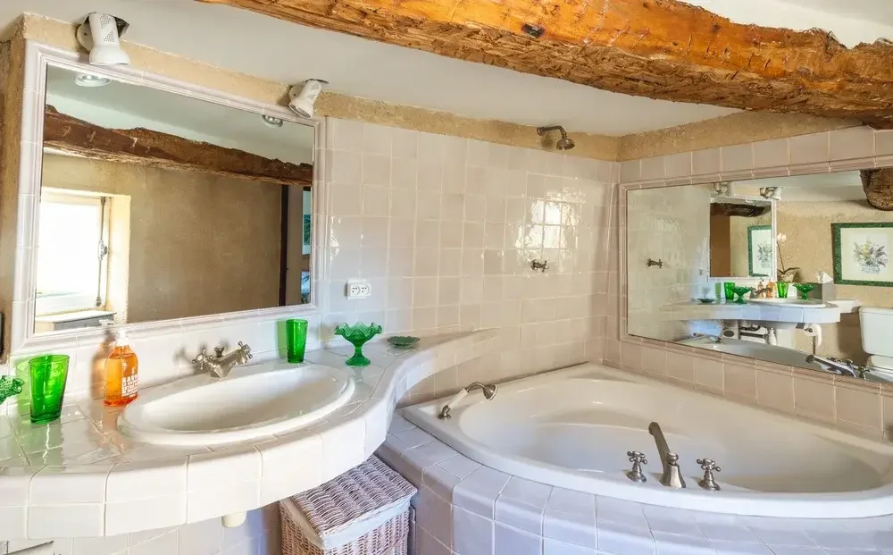Salle de bain avec une baignoire jacuzzi, un lavabo à côté avec un miroir au-dessus, des verres en plastique vert et un savon, murs en carrelage beige, poutre en bois au plafond, fenêtre laissant entrer la lumière naturelle, décoration simple et rust