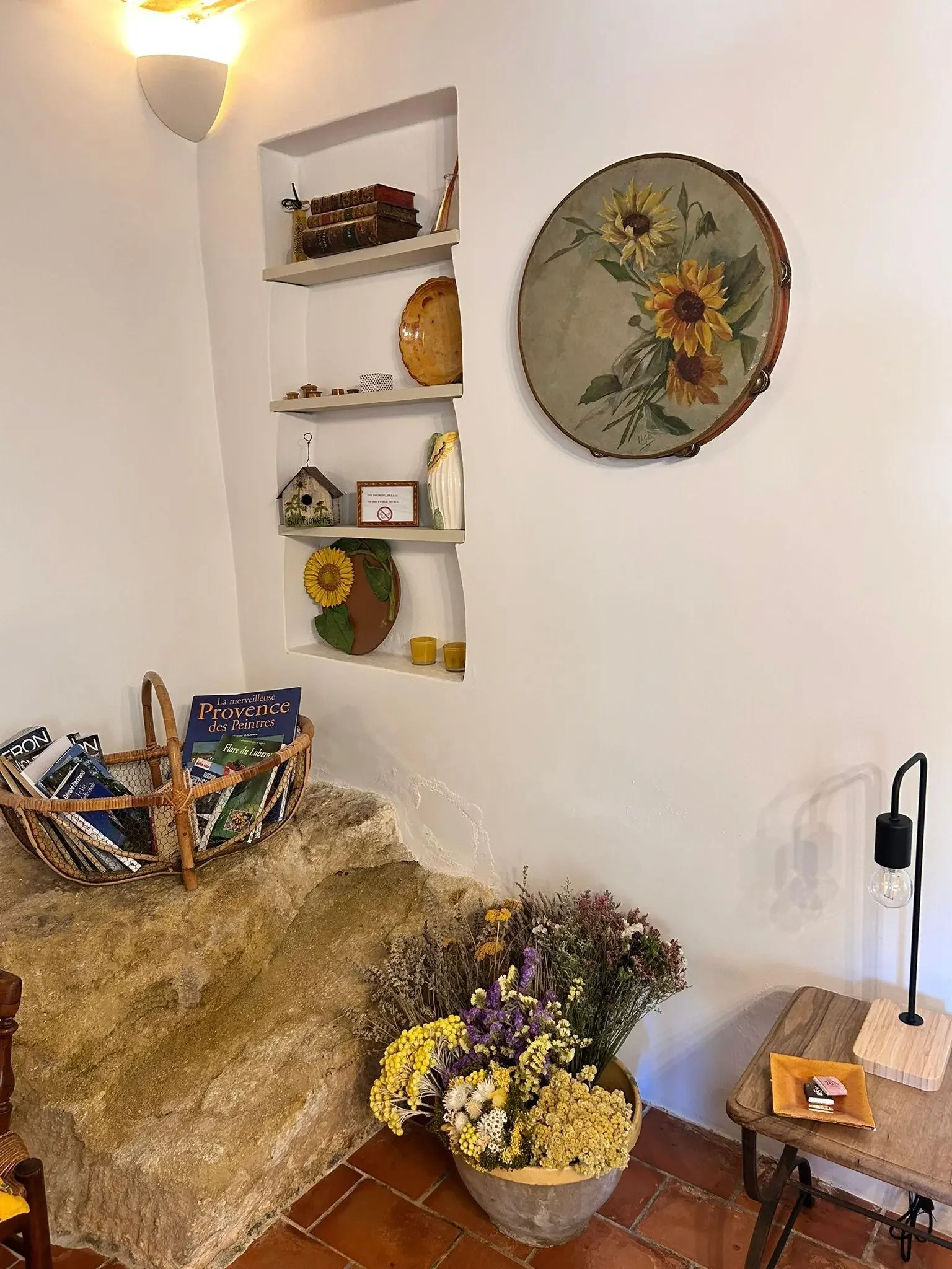 Une étagère blanche encastrée dans un mur blanc, décorée de livres anciens, de céramiques, d'une plaque avec un message, et une décoration en bois représentant une tête de tournesol. Au dessous, un pot de fleurs avec des fleurs jaunes, blanches, et violet, posé sur un sol en terre cuite. À côté, un petit meuble en bois avec un plateau en céramique et une lampe à ampoule nue.
