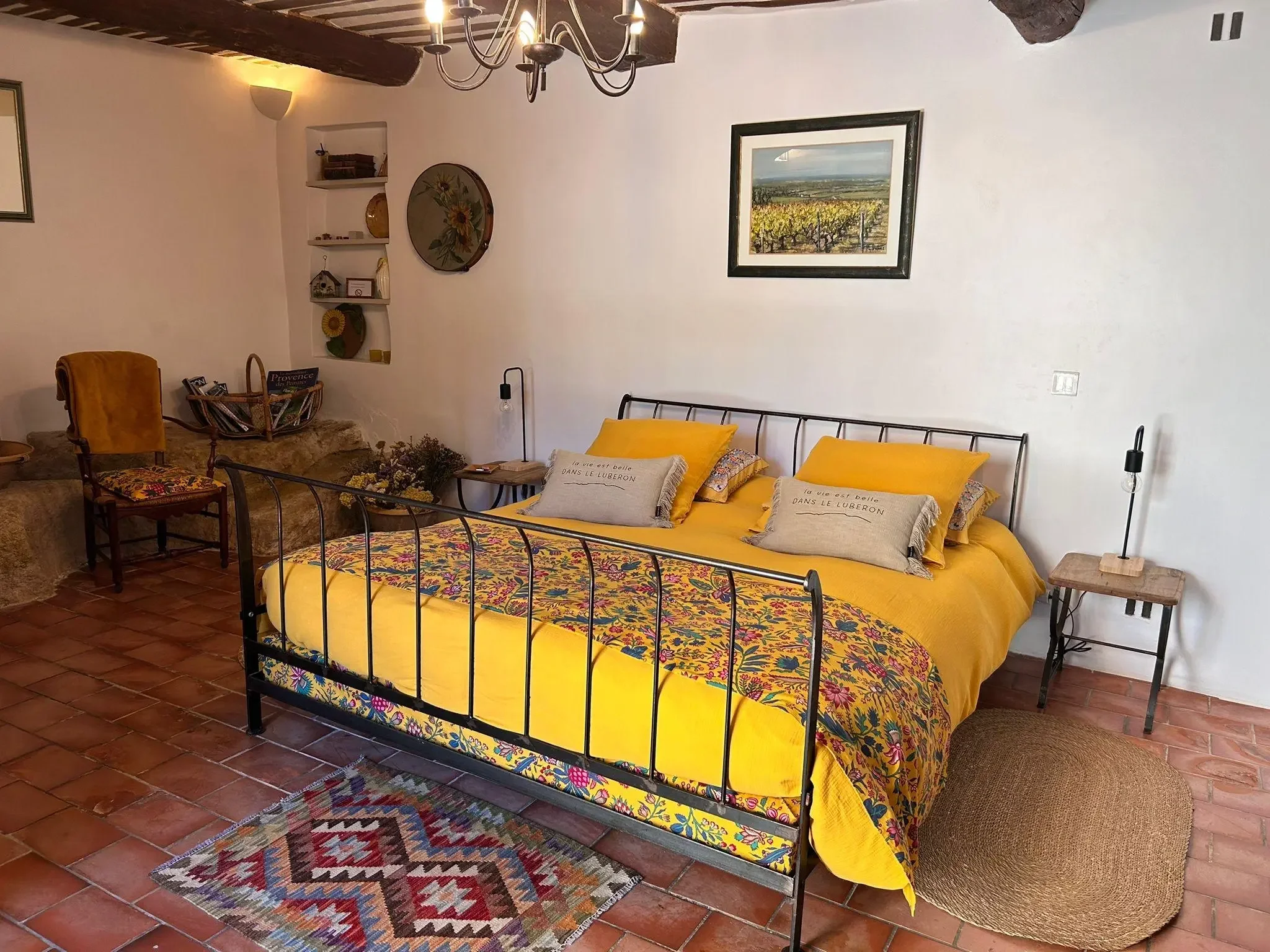Une chambre de style rustique avec un lit en métal noir, des draps jaune vif et des coussins avec des citations en français. Il y a une petite table en bois avec une lampe, une chaise en bois avec un coussin coloré, un tapis en laine multicolore et des décorations murales, dont un tableau d'un paysage viticole et des étagères avec des objets décoratifs.