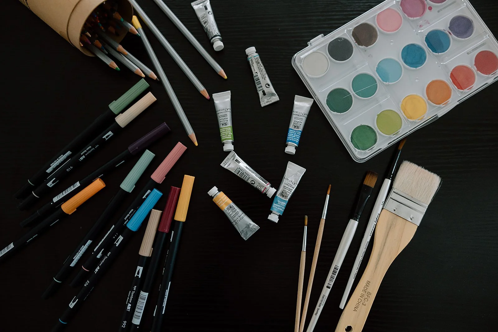 A collection of art supplies on a black surface, including watercolor paints, acrylic paints, colored markers, brushes, and colored pencils.