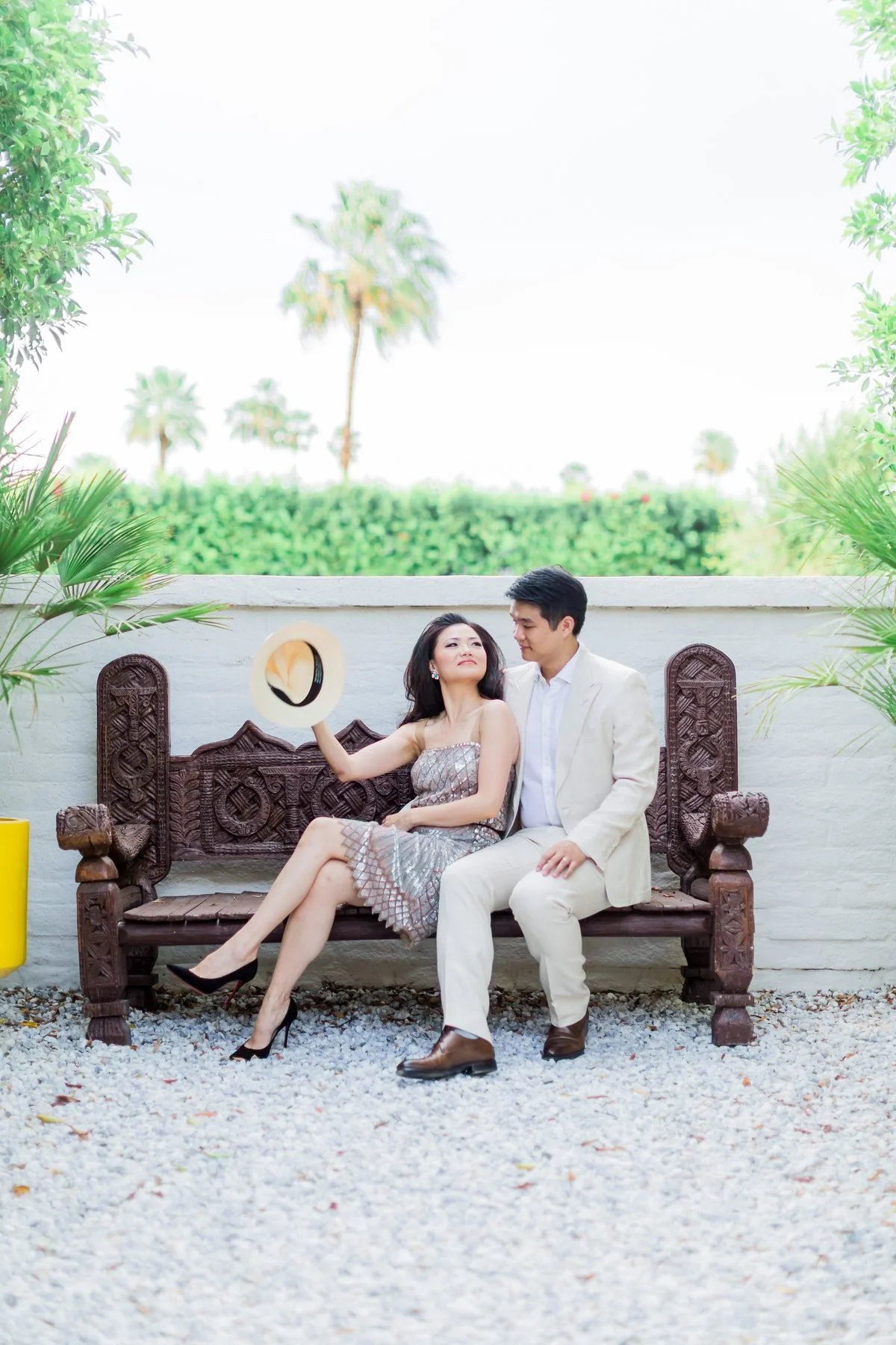 Natalie holding her sun hat against the breeze while sitting close to her husband on a carved wooden bench in the Palm Springs garden.