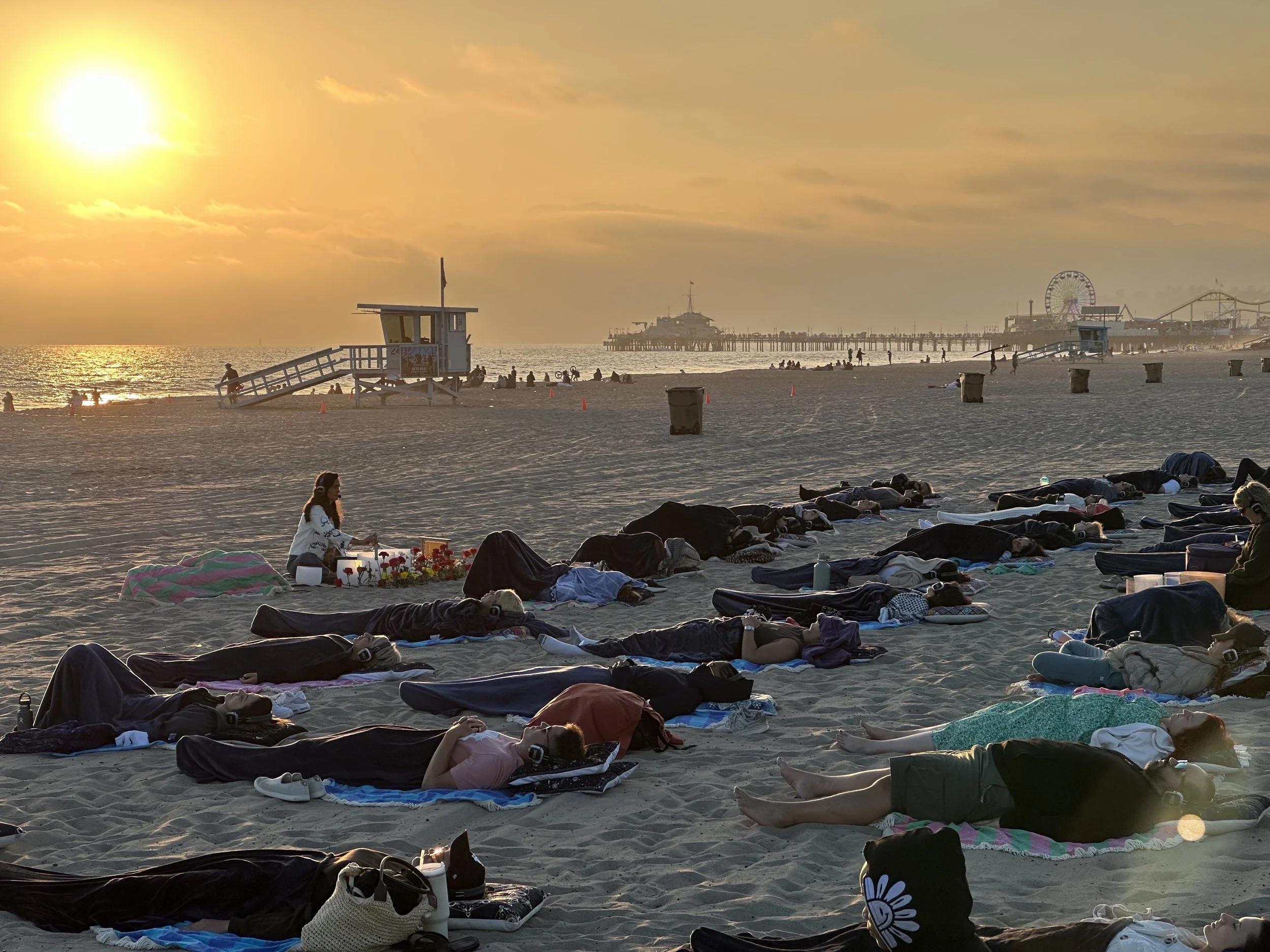 People sitting on mats and blankets on the beach, watching the sunset over the ocean with clouds in the sky.