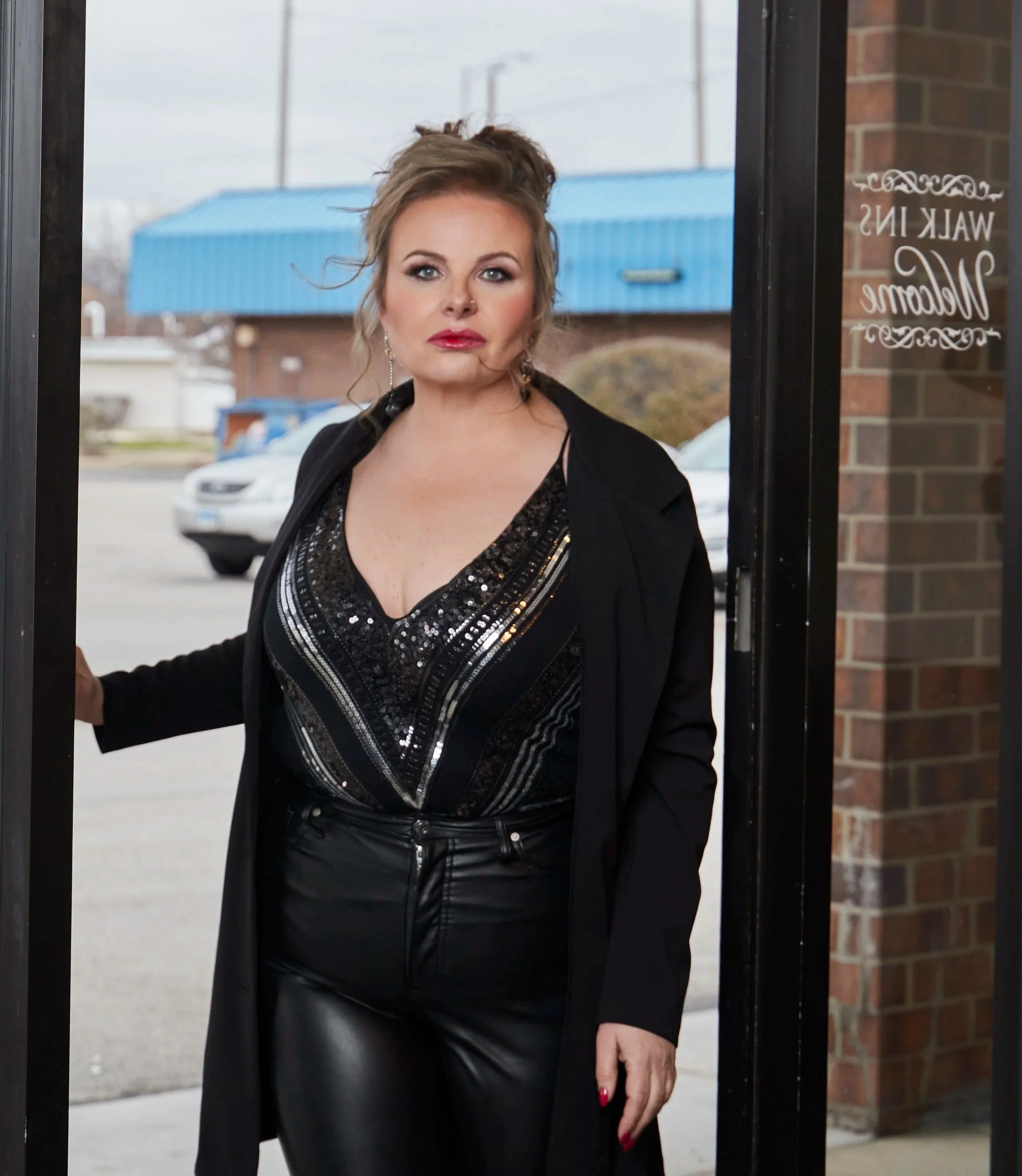 A woman with curly, light brown hair and red lipstick standing at a doorway. She is wearing a black outfit with sequined details and leather pants, with earrings and a nose ring. Behind her, a street with parked cars, a brick building, and a blue industrial building are visible. The door has a sign that says 'WALK IN Welcome' in decorative writing.