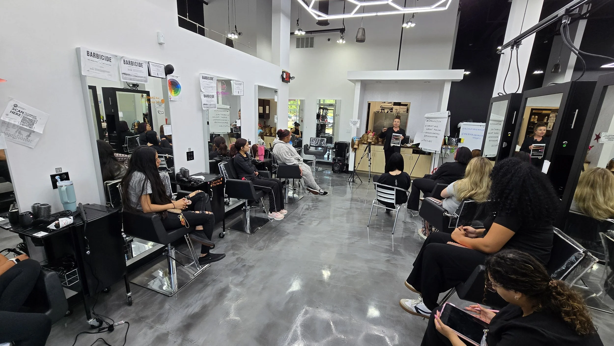 A classroom or training room filled with women seated in chairs, facing a speaker at the front, with hair and makeup tools visible on tables, and whiteboards with notes and posters on the walls.