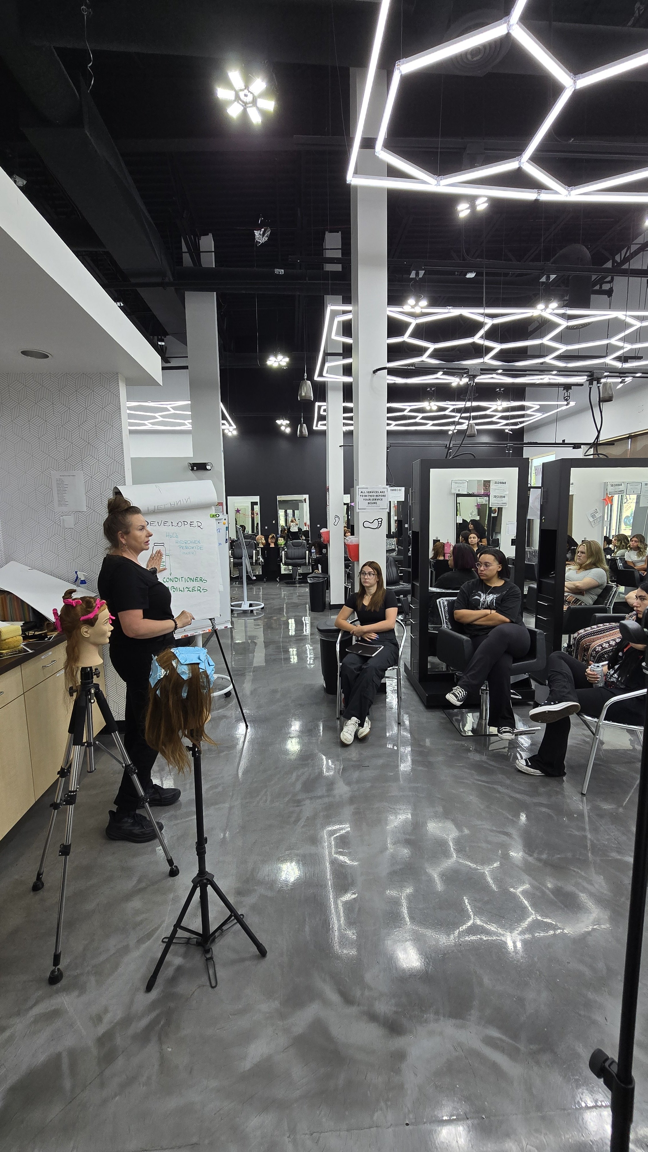 A woman is standing and giving a presentation in a hair salon classroom, with hair mannequins and students seated in chairs watching her.