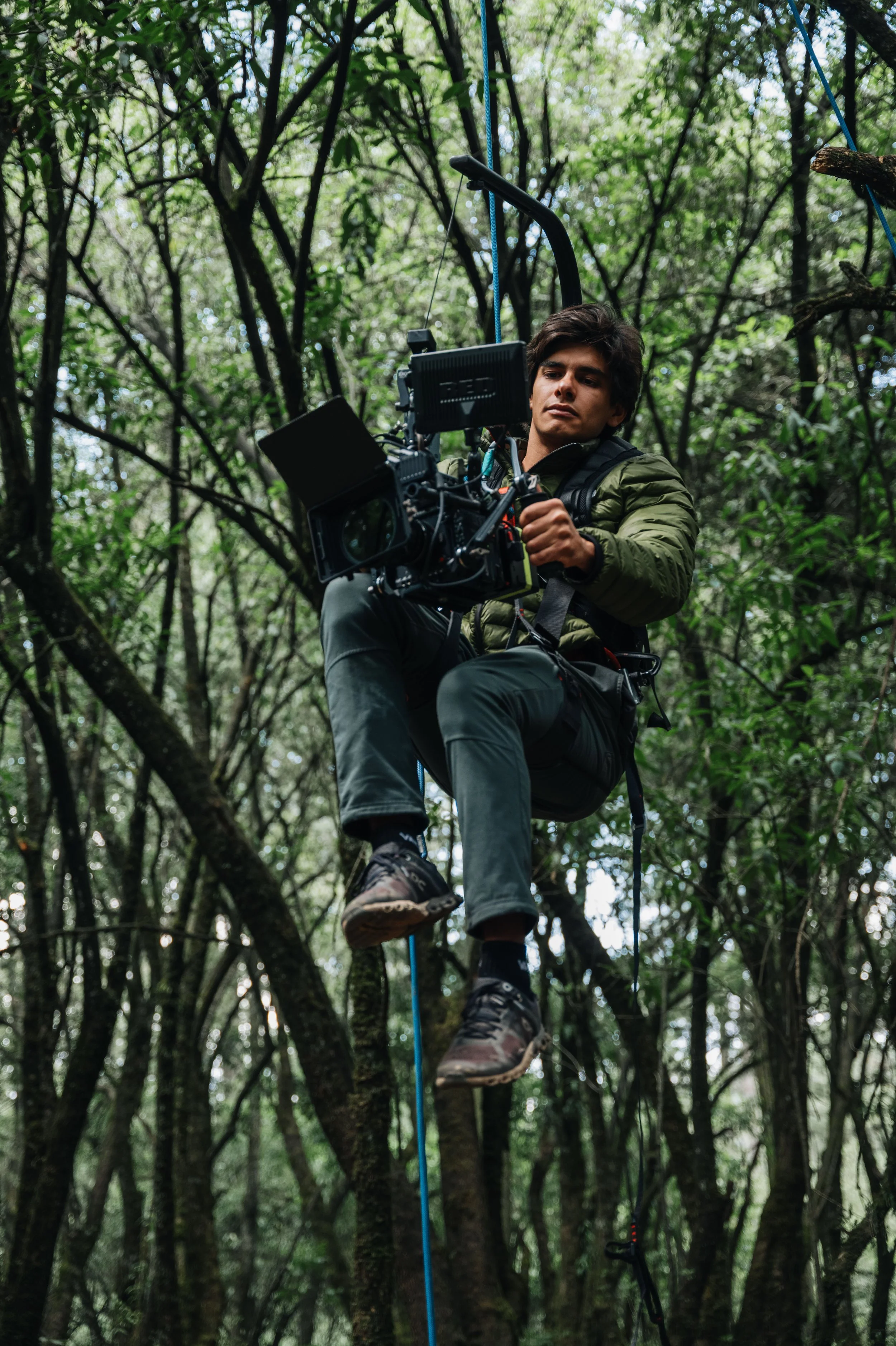 Hombre en arnés, caminando sobre cuerda en un bosque, usando equipo para escalada o tirolesa, rodeado de árboles verdes.