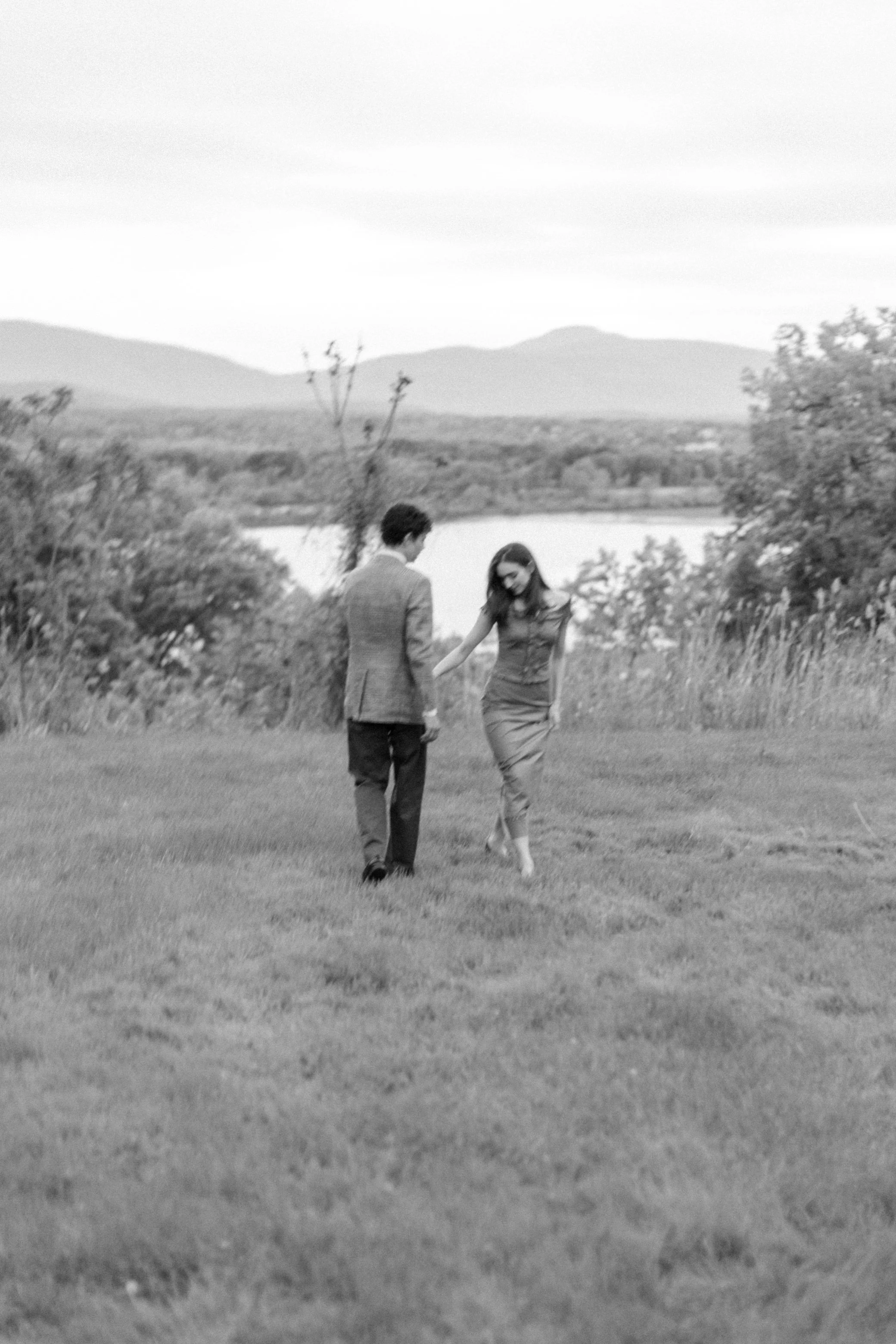 black and white photograph of engaged couple walking in the grass with the hudson river and catskill mountains in the background