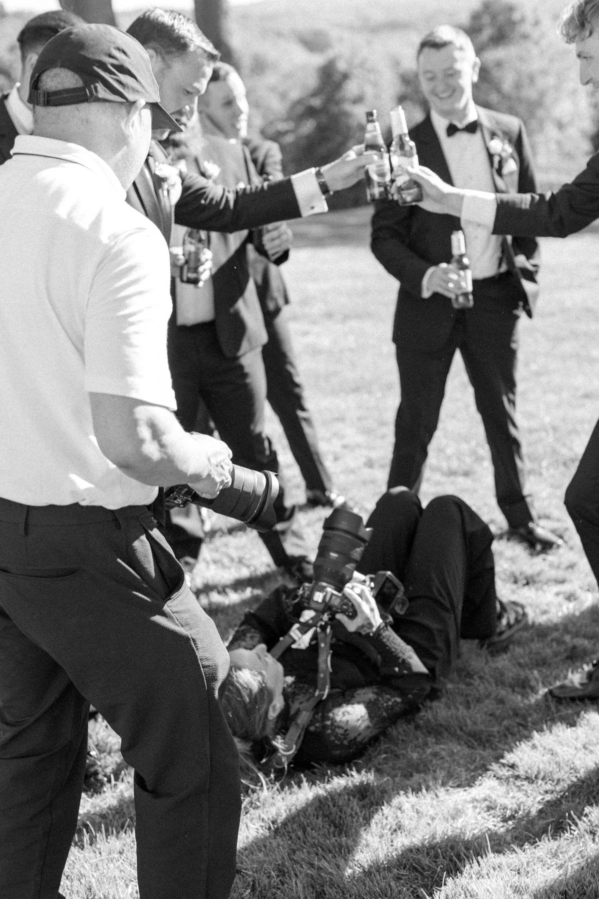 Groomsmen toasting at a wedding outdoors, with Caitlinn Mahar-Daniels, hudson valley wedding photographer, lying on the grass taking photographs.