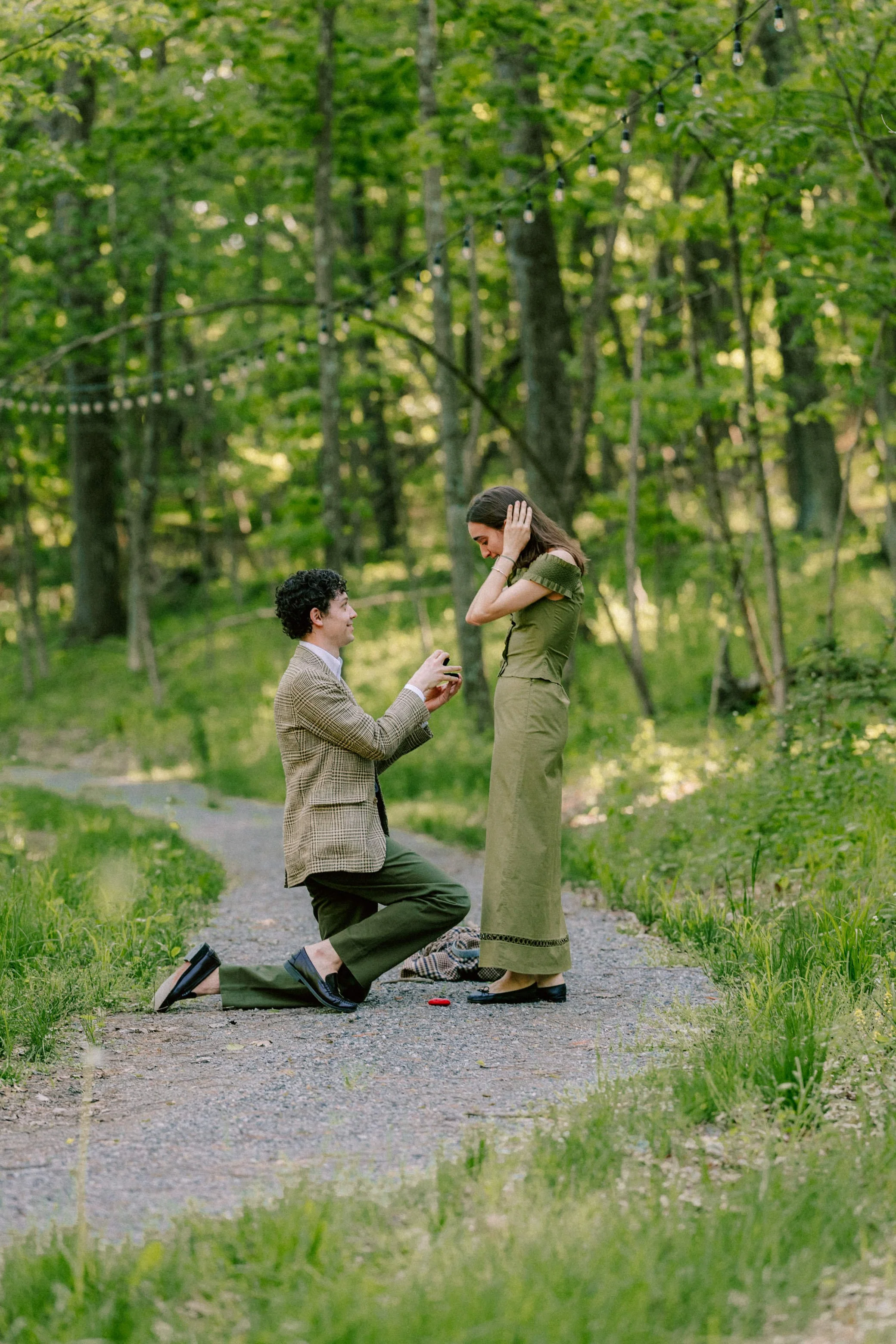 man is on one knee proposing to woman in green dress on a path in the woods at camptown in the catskills
