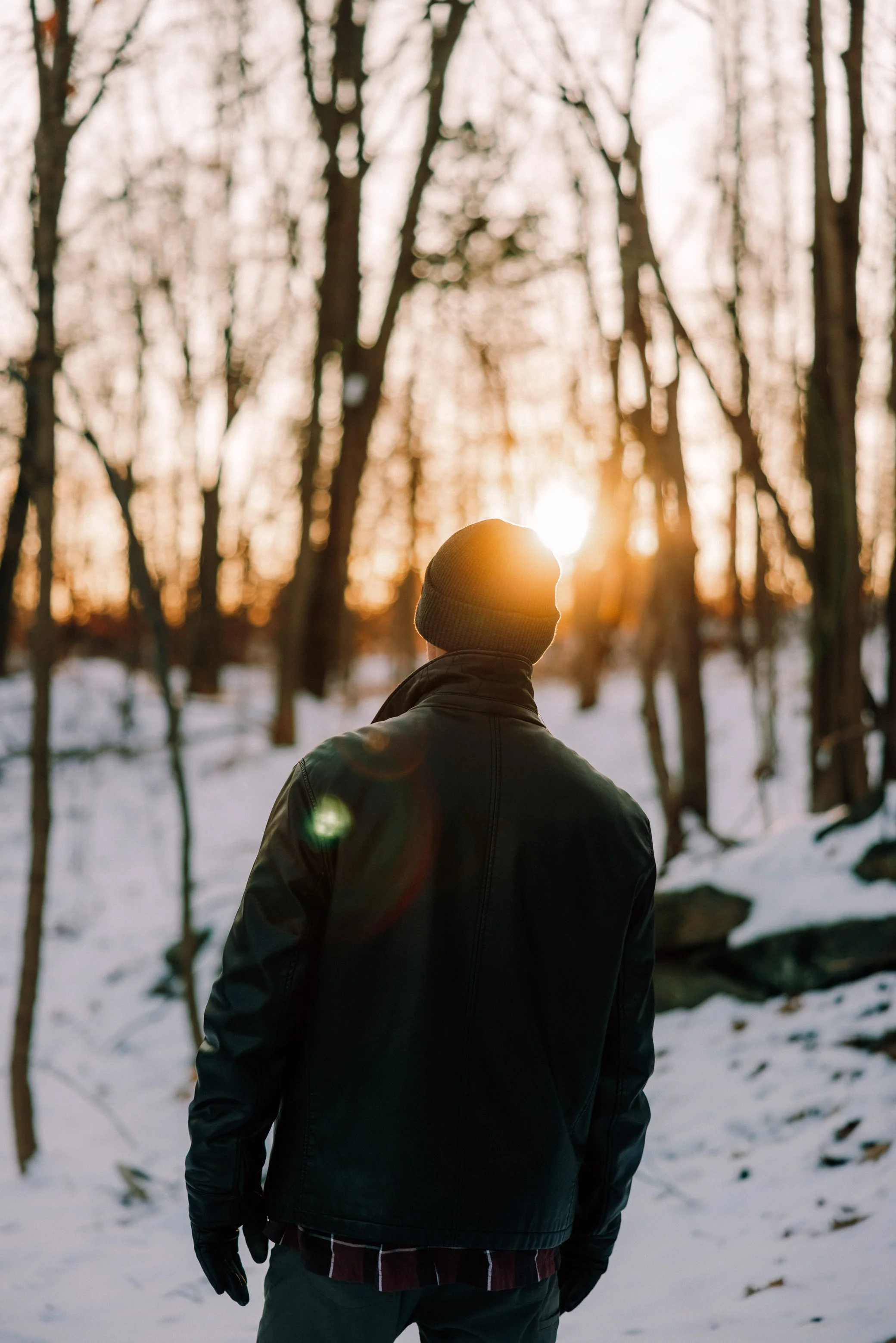 Caitlinn Mahar-Daniels' husband dressed in dark winter clothing, including a beanie and gloves, walking through a snow-covered forest at sunset in the Hudson Valley.