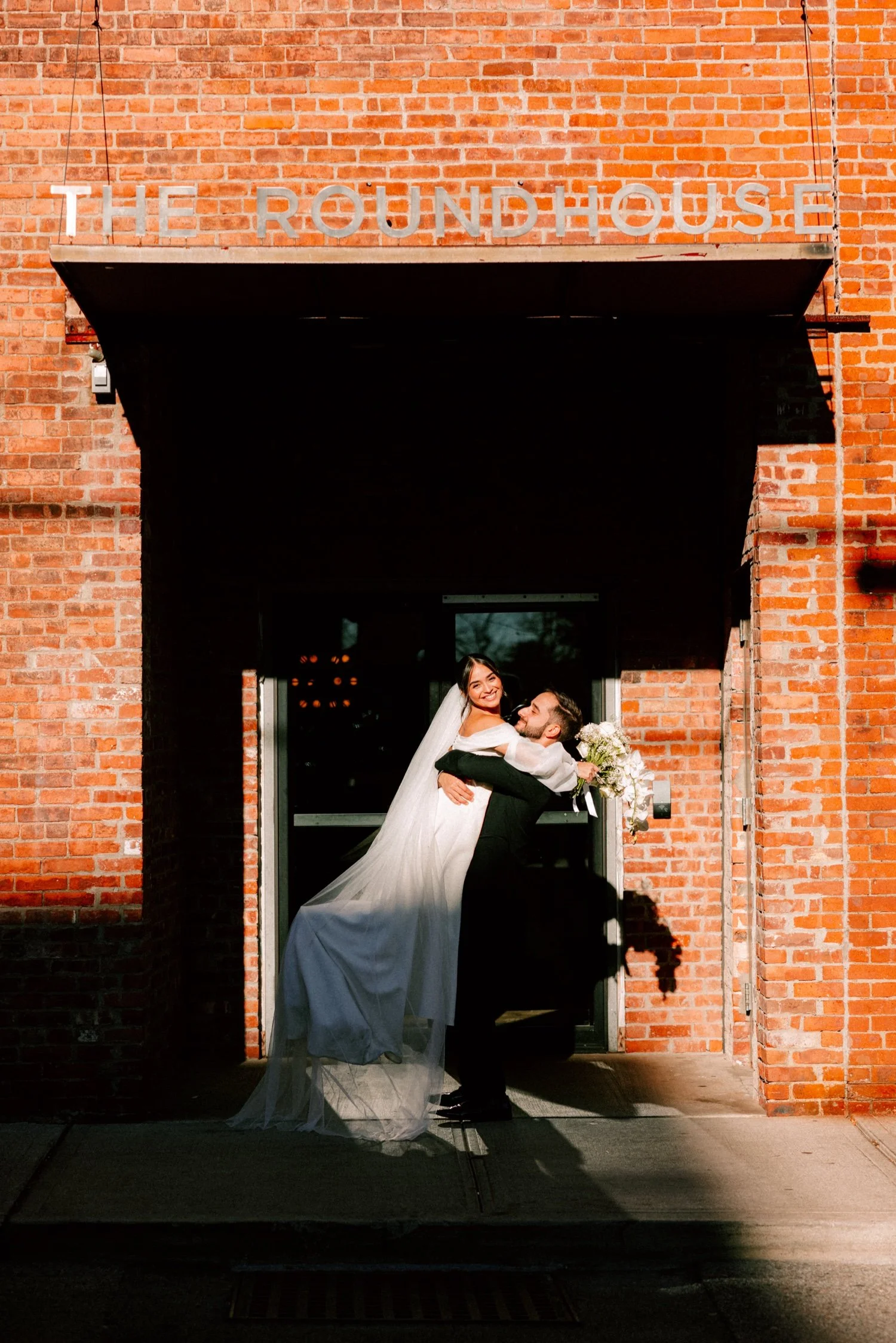groom lifts bride while she looks at the camera and smiles in the direct sun on their wedding day at the roundhouse in beacon ny