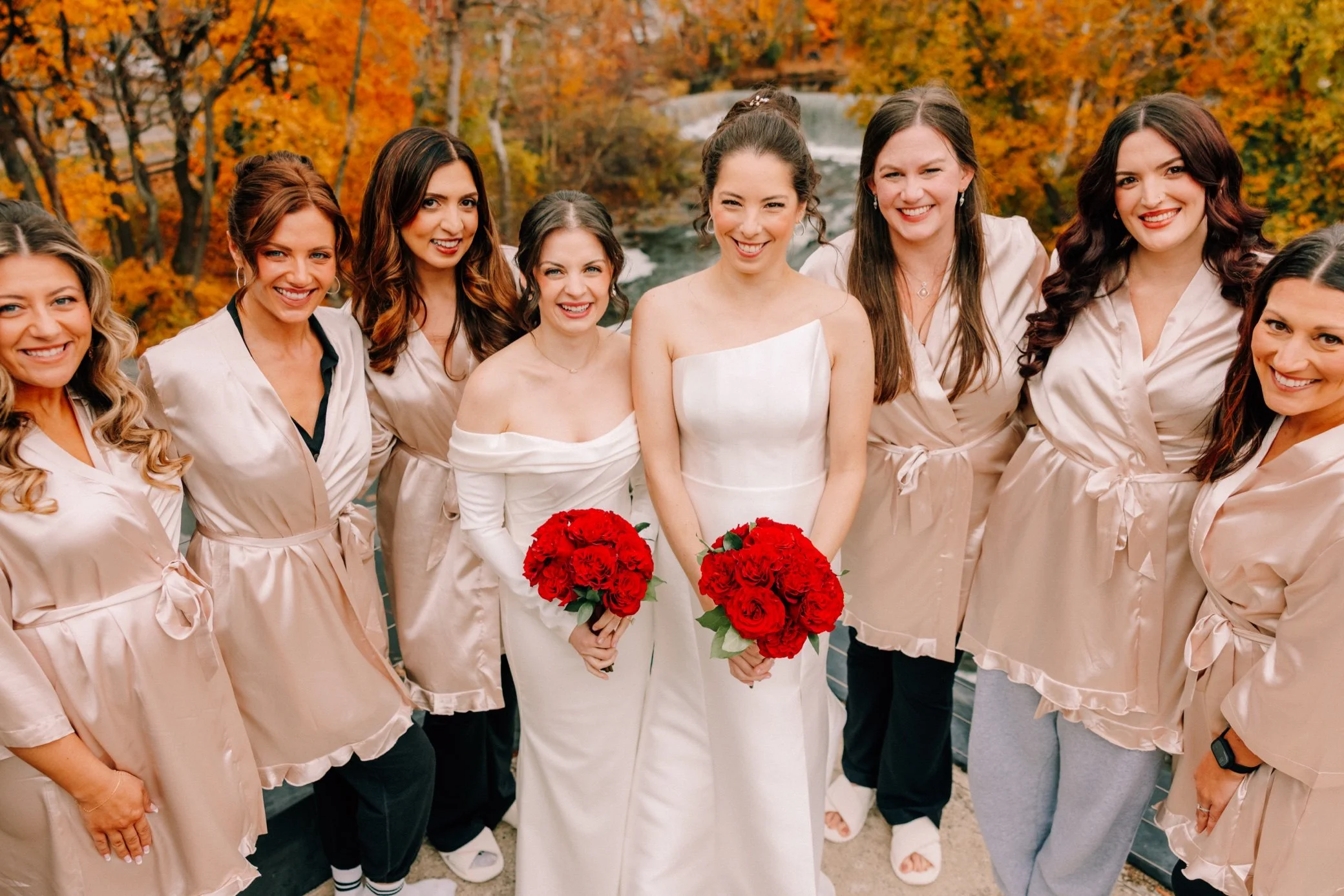  brides with their wedding dresses holding red bouquets with their bridesmaids in robes on the penthouse balcony with beacon falls in the background at the roundhouse 