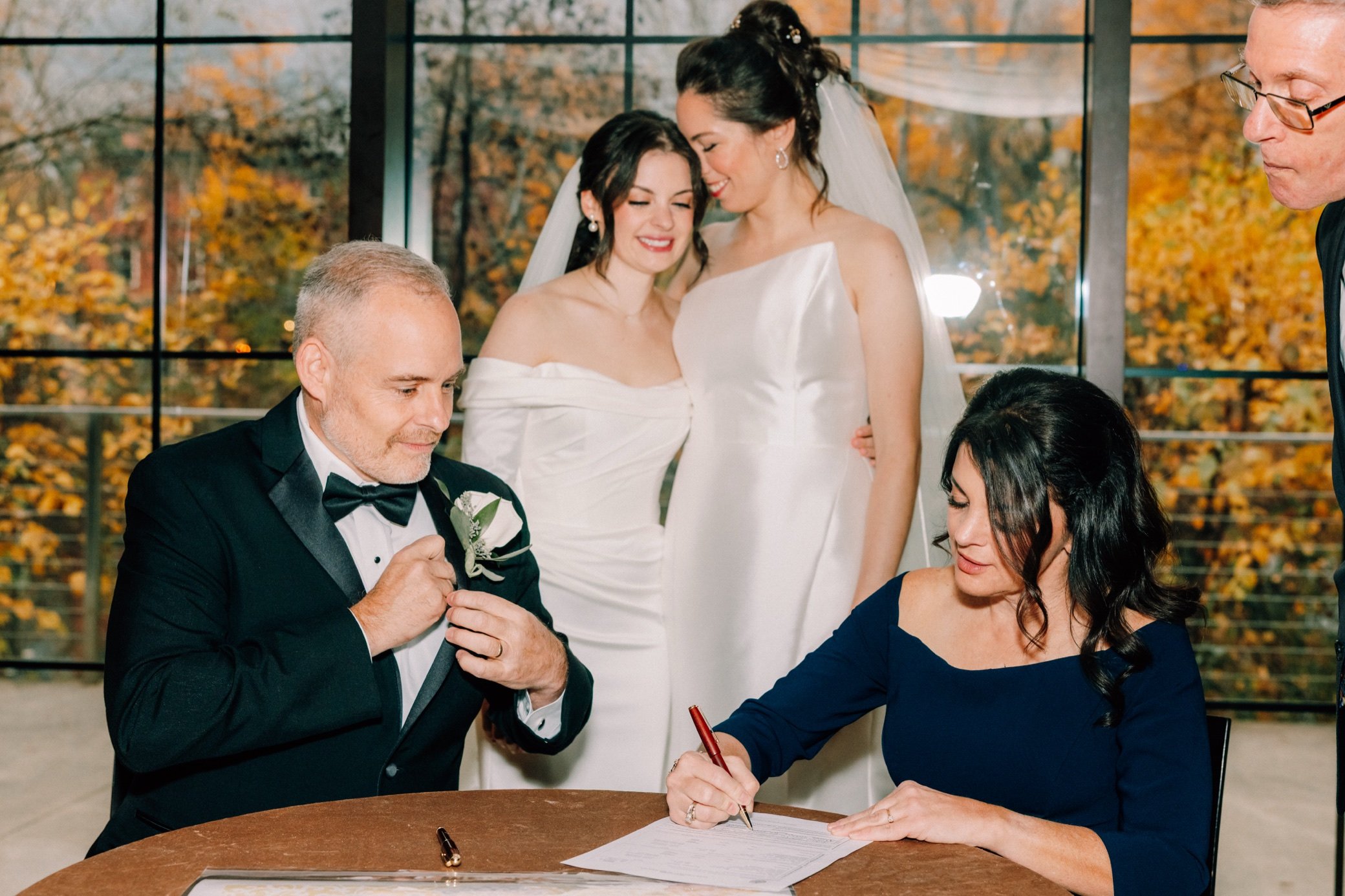  bride’s parents sign the ketubah while brides embrace while behind them at the roundhouse 