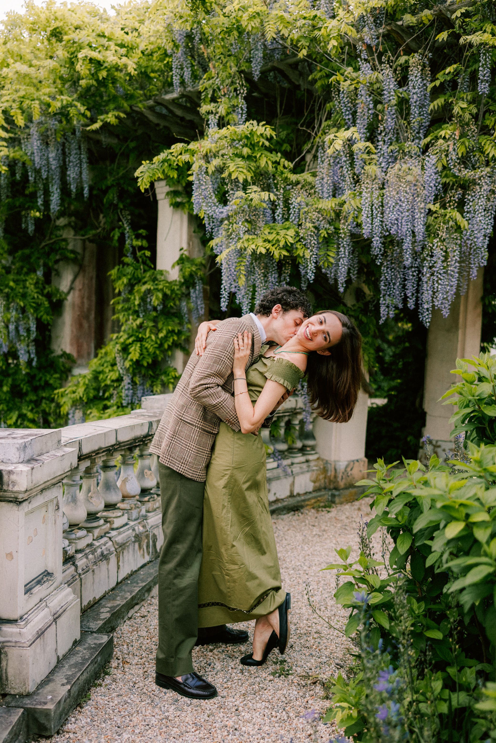 engaged couple embrace in a garden while he kisses her on her neck with wisteria behind them