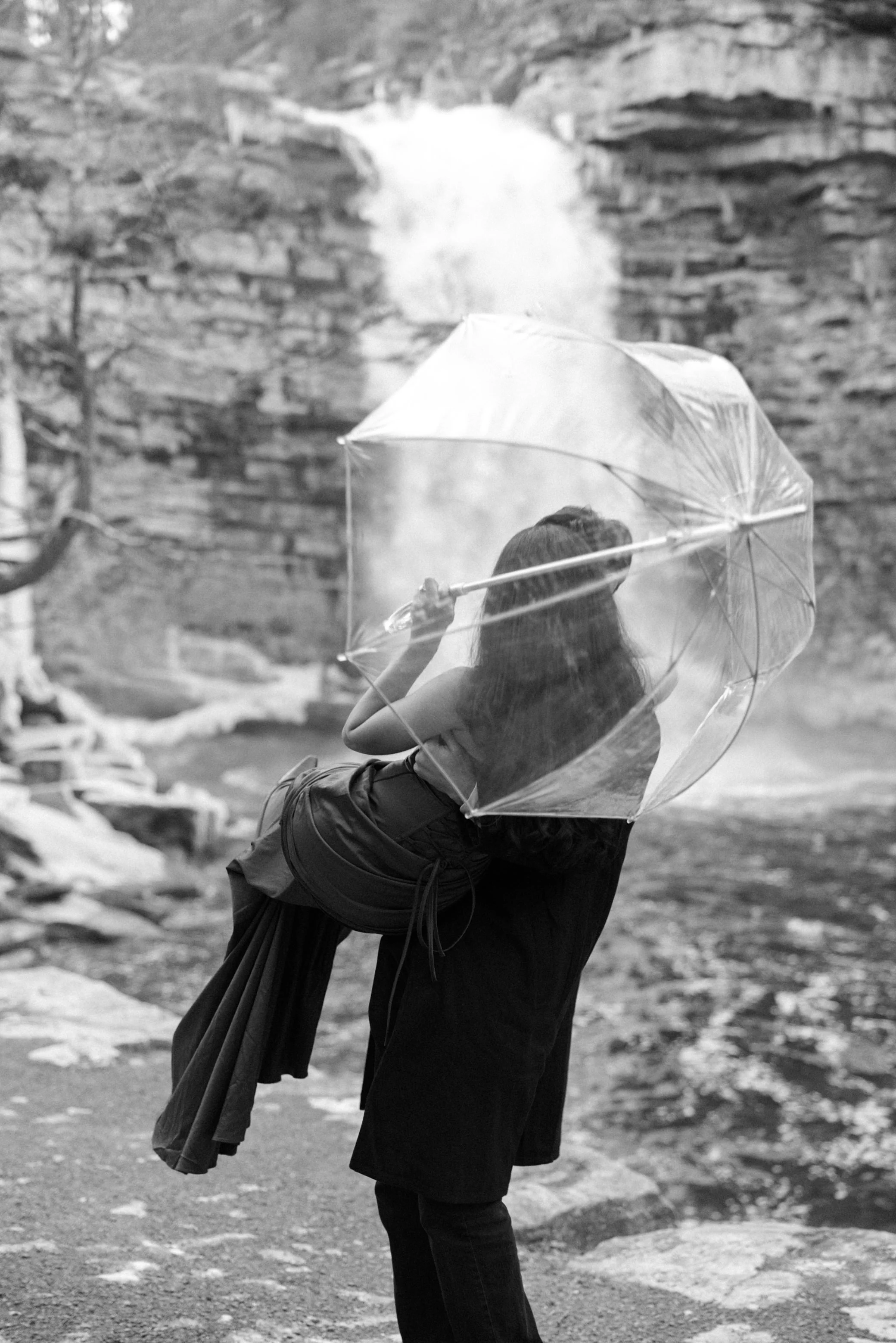 A woman being held by her new fiance holding a transparent umbrella during their engagement shoot at Awosting Falls after their surprise proposal at Mohonk Mountain House.