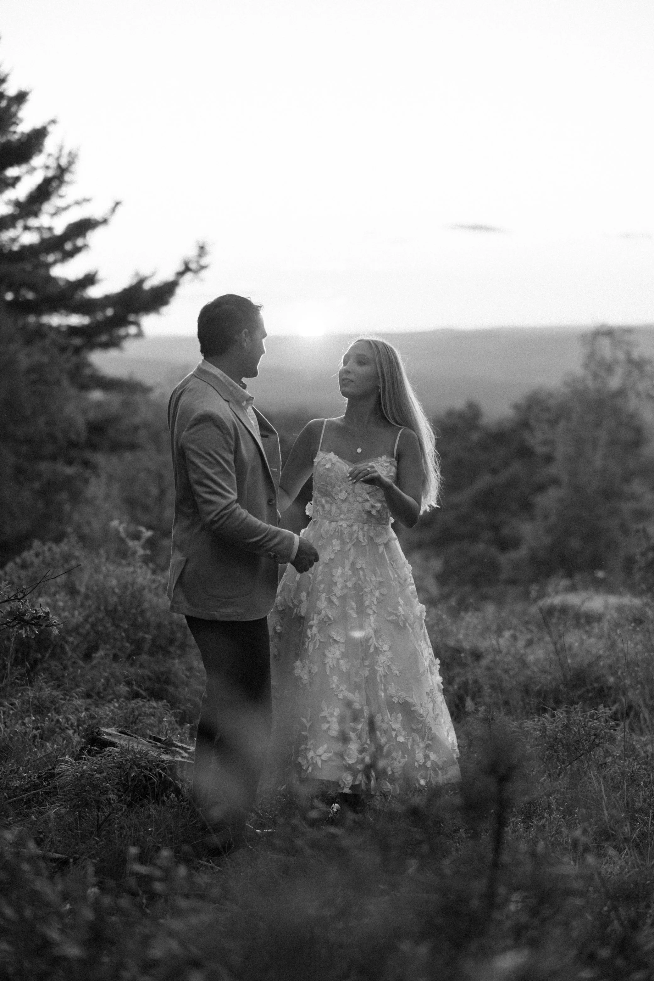 A black and white photo of a couple standing outdoors on a hill at sunset, with a scenic landscape in the background. The woman wears a floral lace dress and the man wears a blazer. They are looking at each other.