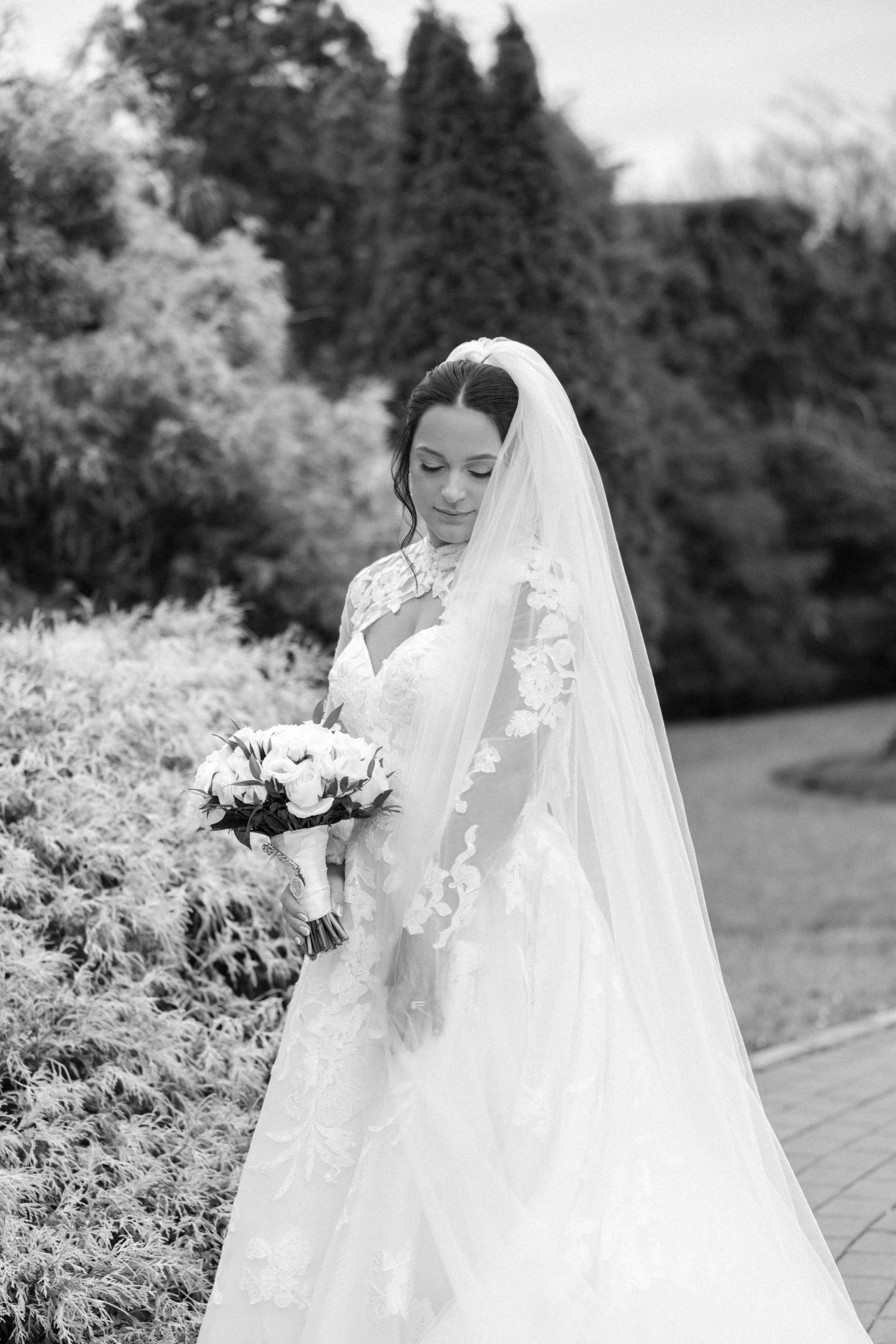 bride in lace long sleeve wedding dress with cathedral length veil stands in a garden in winter on her hudson valley wedding day at VIP Country Club