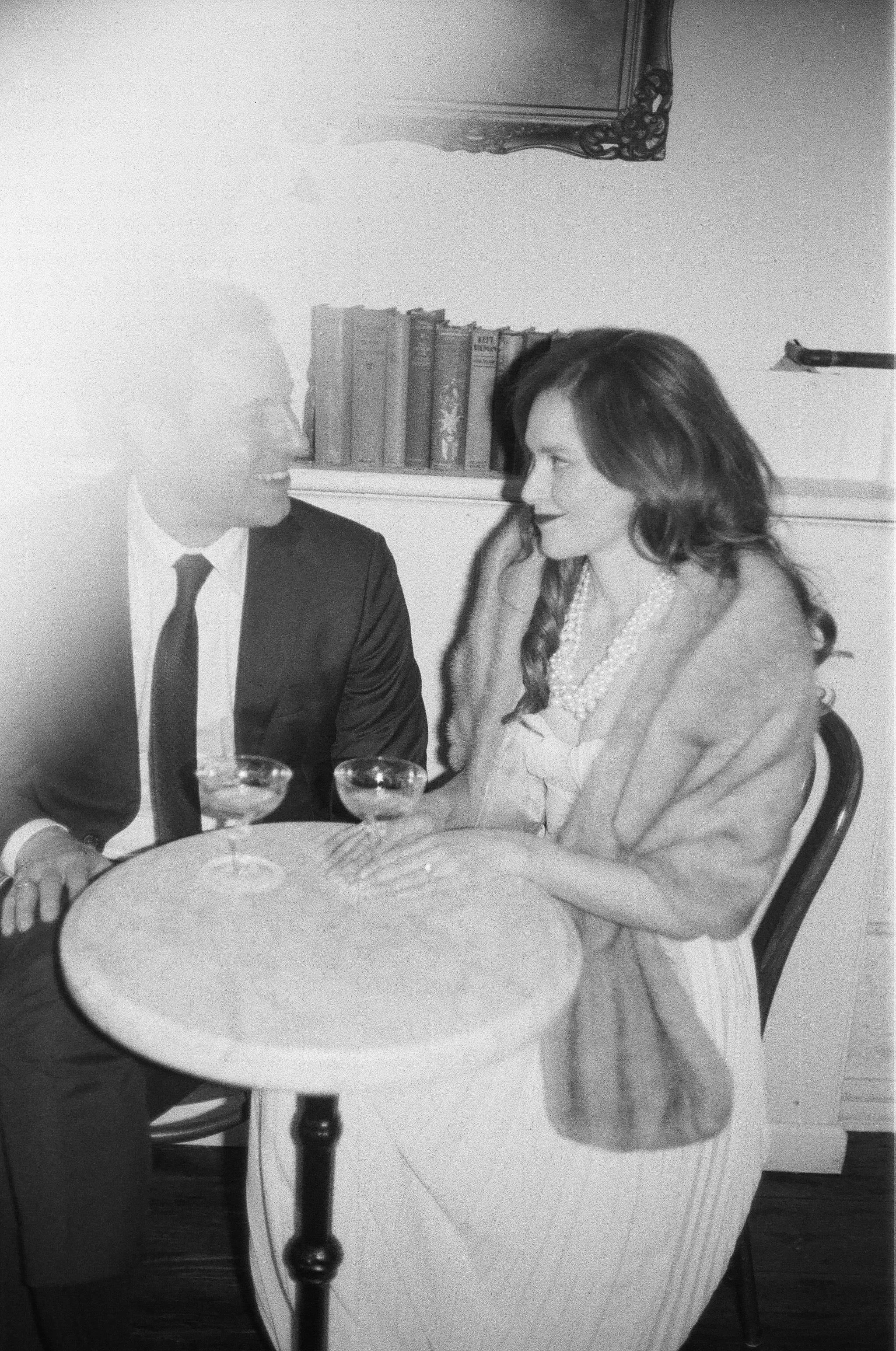 black and white film photograph of engaged couple sitting at bistro table with coupe glasses on it, wearing old hollywood inspired clothing for their engagement photo shoot at brunette wine bar in kingston new york