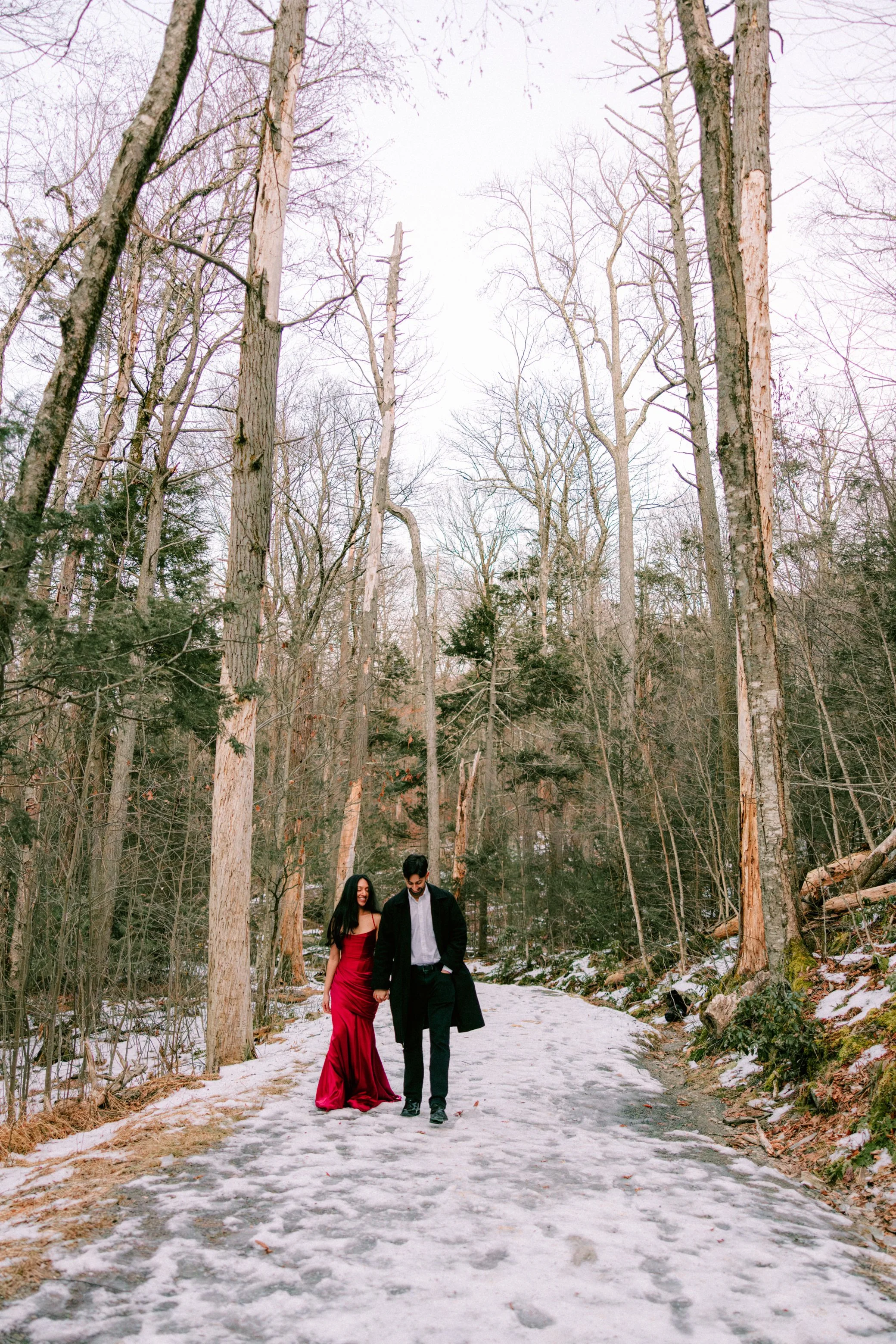 A couple walking hand in hand on a snowy forest path during winter in Minnewaska State Park. The woman is wearing a red gown and the man is dressed in dark clothing.