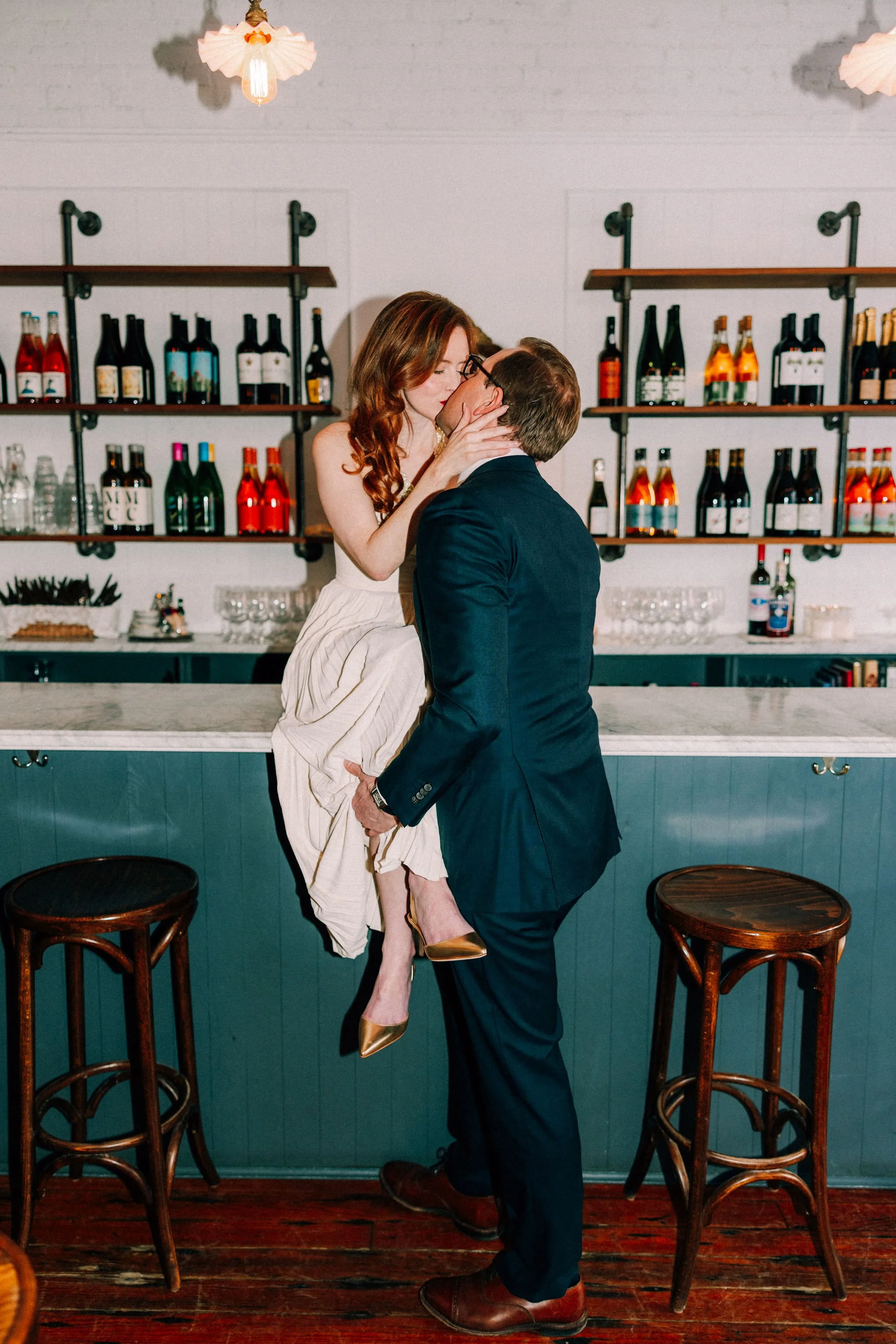 woman sits on bar wearing white dress and golden high heels holding onto mans face while they kiss during their vintage inspired engagement photography shoot in brunette wine bar in kingston new york