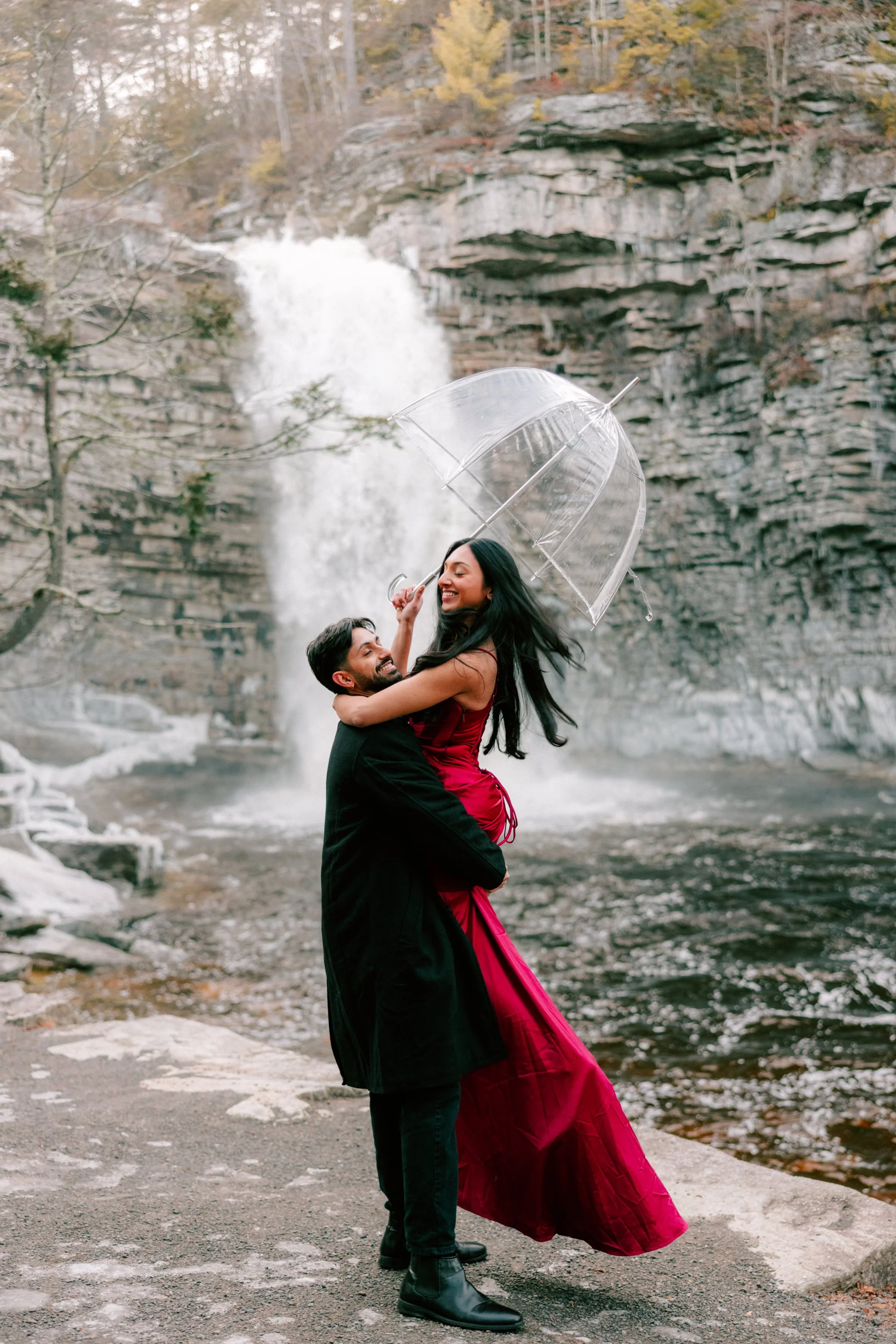 A smiling couple, with the woman in a long red dress, holding a transparent umbrella, standing in front of Awosting Falls in winter during their hudson valley engagement photography shoot.