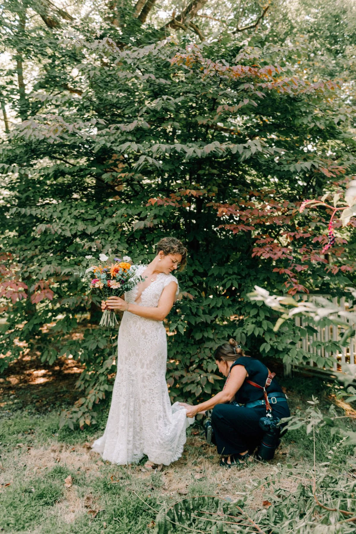 Behind the scenes photograph of a woman in a white lace wedding dress holding a bouquet of colorful flowers is having her shoe adjusted by Caitlinn Mahar-Daniels, hudson valley wedding photographer, at The Yard in Beacon, NY.