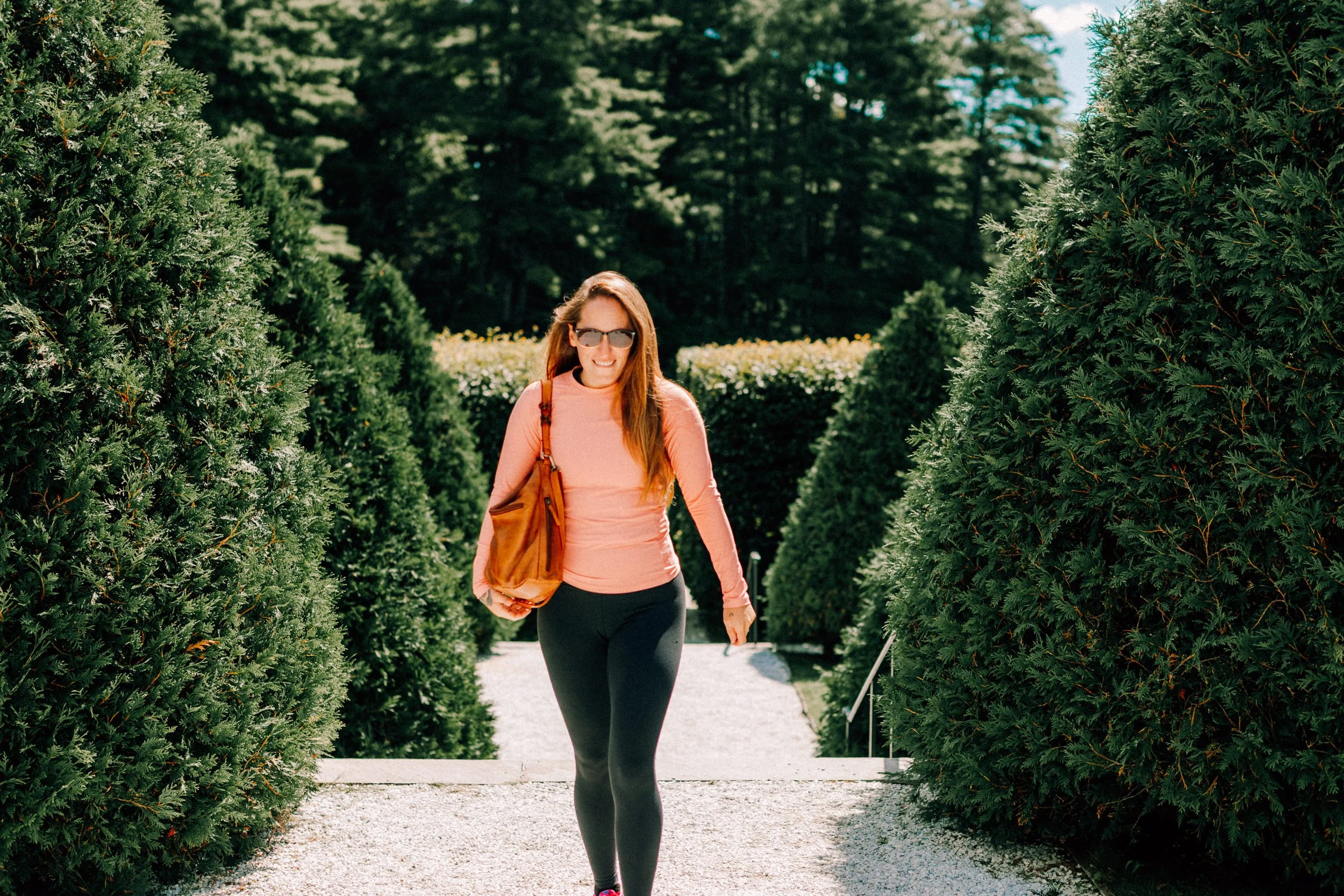 Caitlinn Mahar-Daniels, hudson valley wedding photographer, wearing sunglasses, a pink long-sleeve shirt, and black leggings walking outdoors along a gravel path, surrounded by tall green bushes and trees on a sunny day at The Mount in The Berkshires