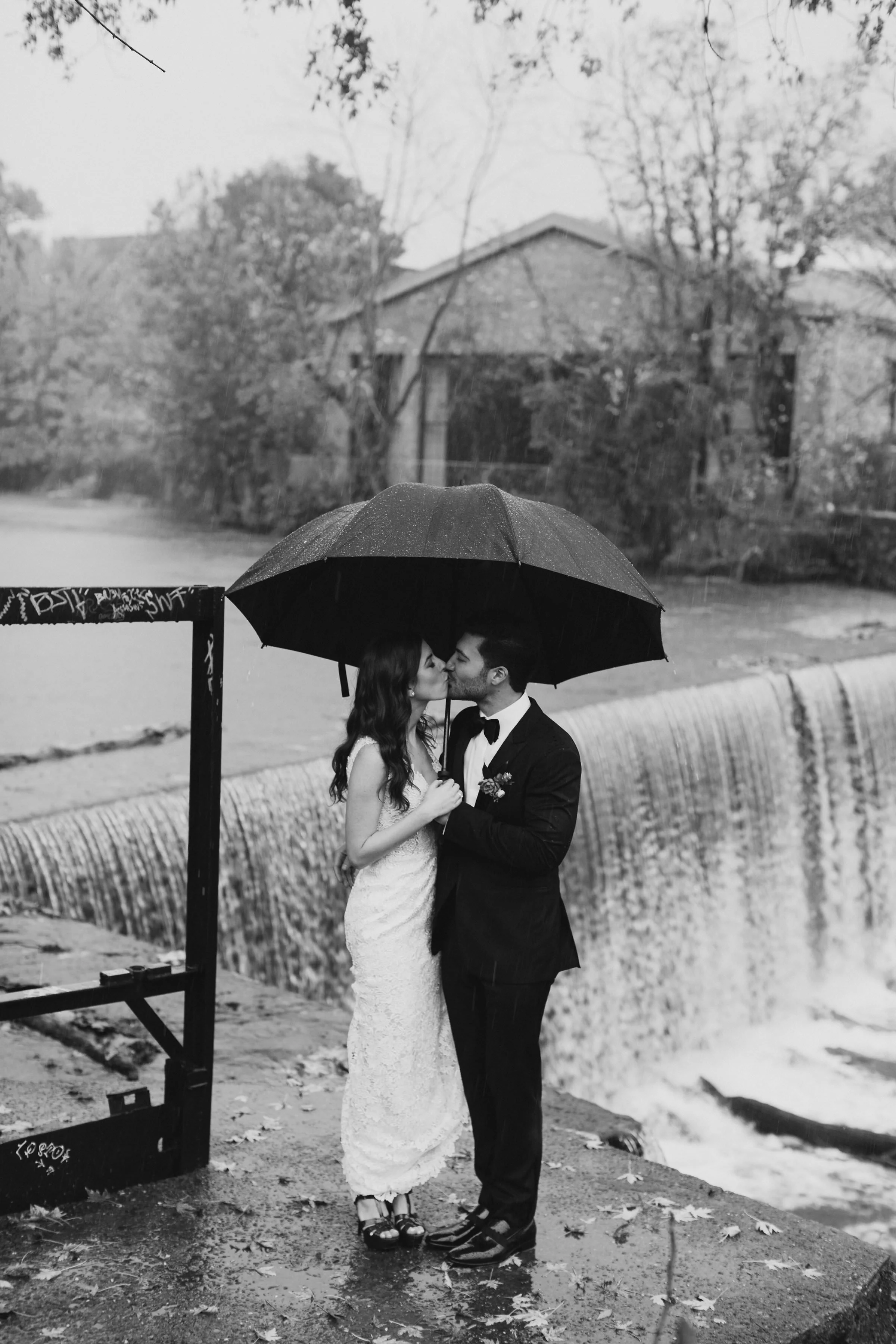 black and white photograph of couple kissing under a black umbrella in the rain in front of Beacon Falls on their wedding day at The Roundhouse