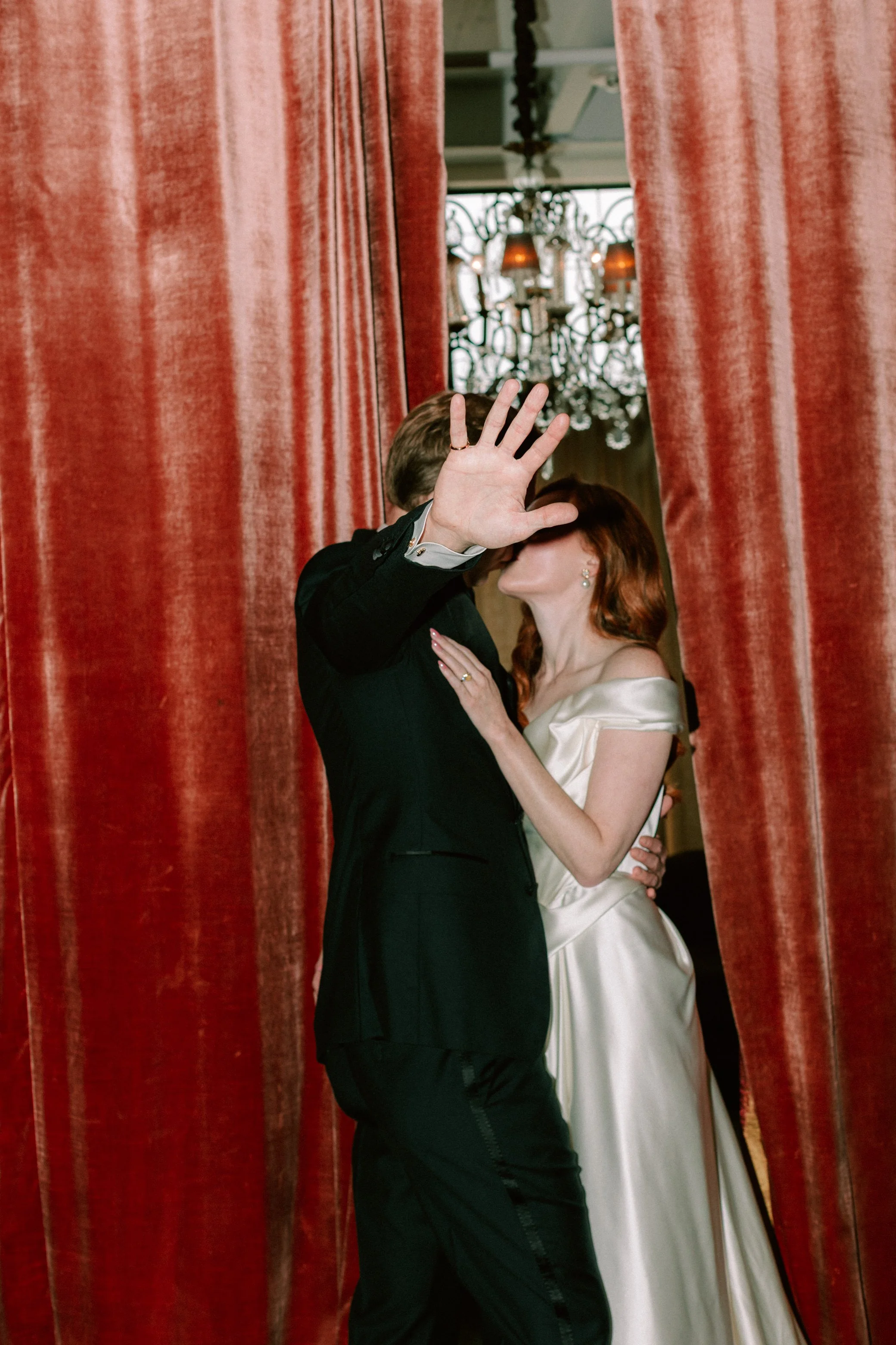 bride and groom kissing in between peach velvet curtains while groom holds up his hand for privacy