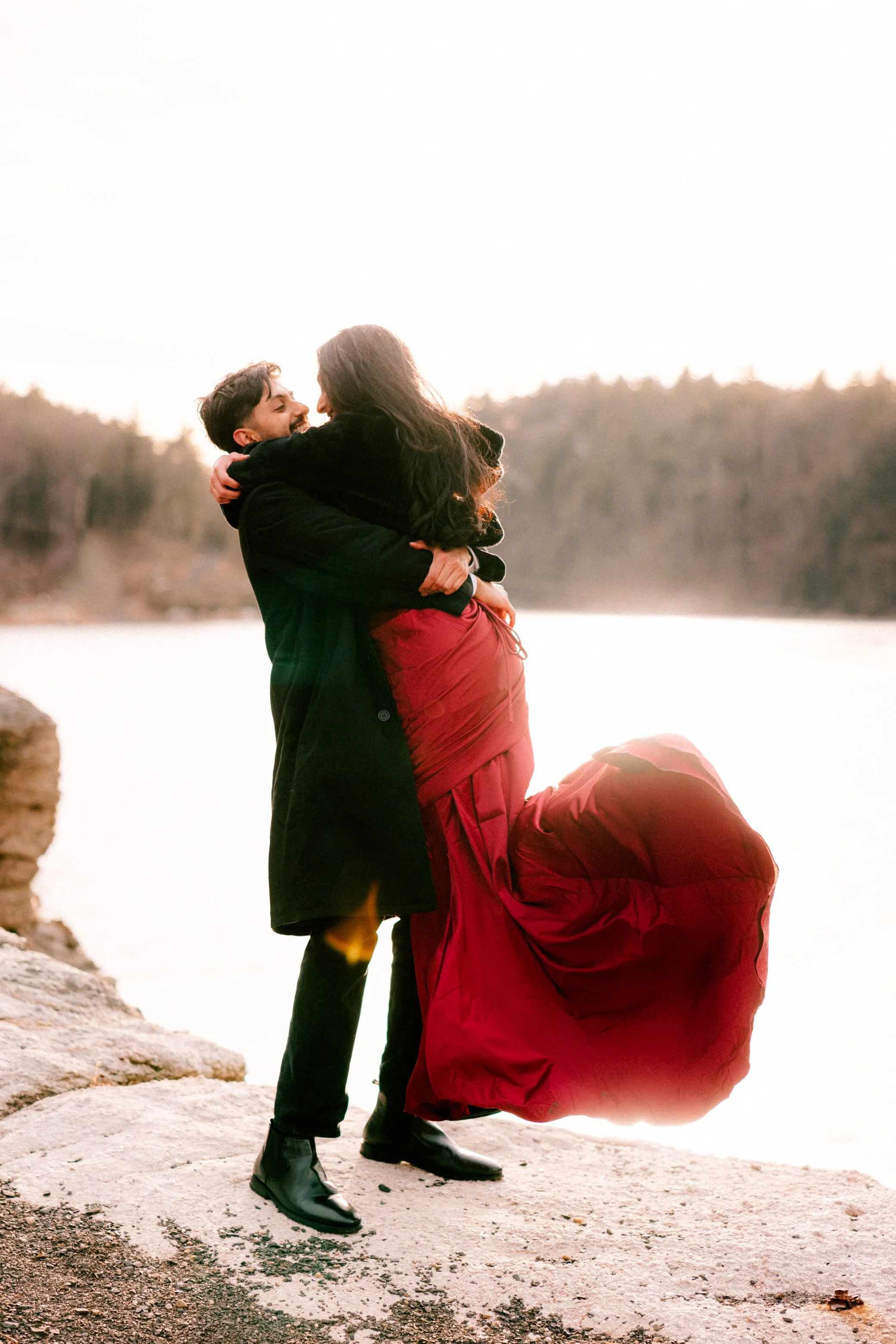 A couple embracing by Lake Mohonk during sunset, with the woman wearing a long red dress and the man in black clothing during their surprise proposal and engagement photo shoot at Mohonk Mountain House.