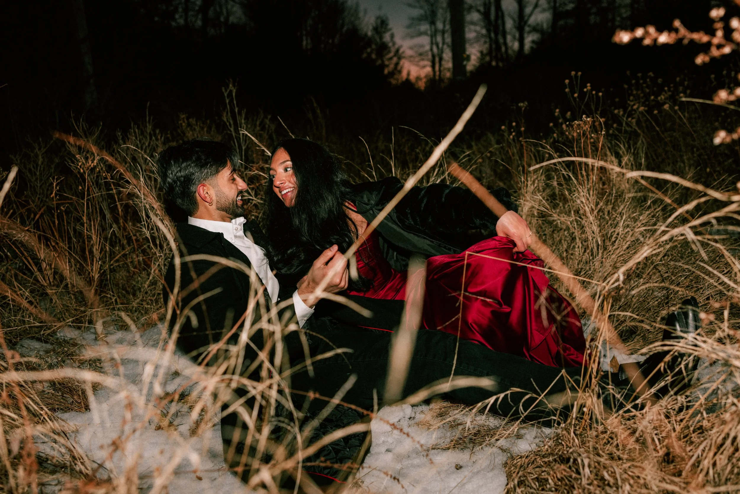 A couple in formal attire lying on ground among tall grass and snow during sunset, smiling and looking at each other during their romantic adventurous engagement photography shoot at Minnewaska State Park.