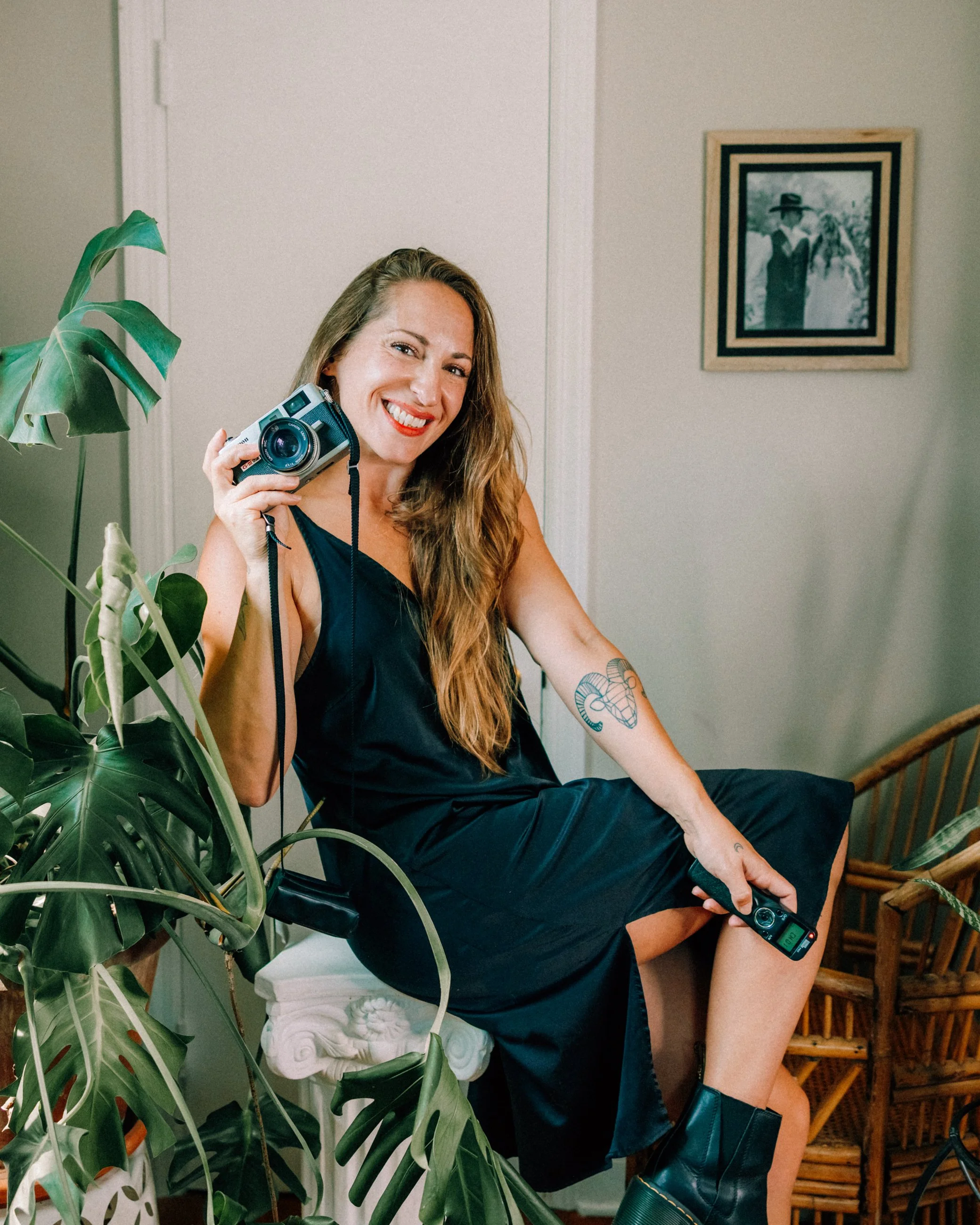 Caitlinn Mahar-Daniels, smiling woman with long wavy hair, wearing a black dress and black boots, sitting on a white column in a room decorated with houseplants. She holds a vintage camera in her right hand and a smartphone in her left hand.