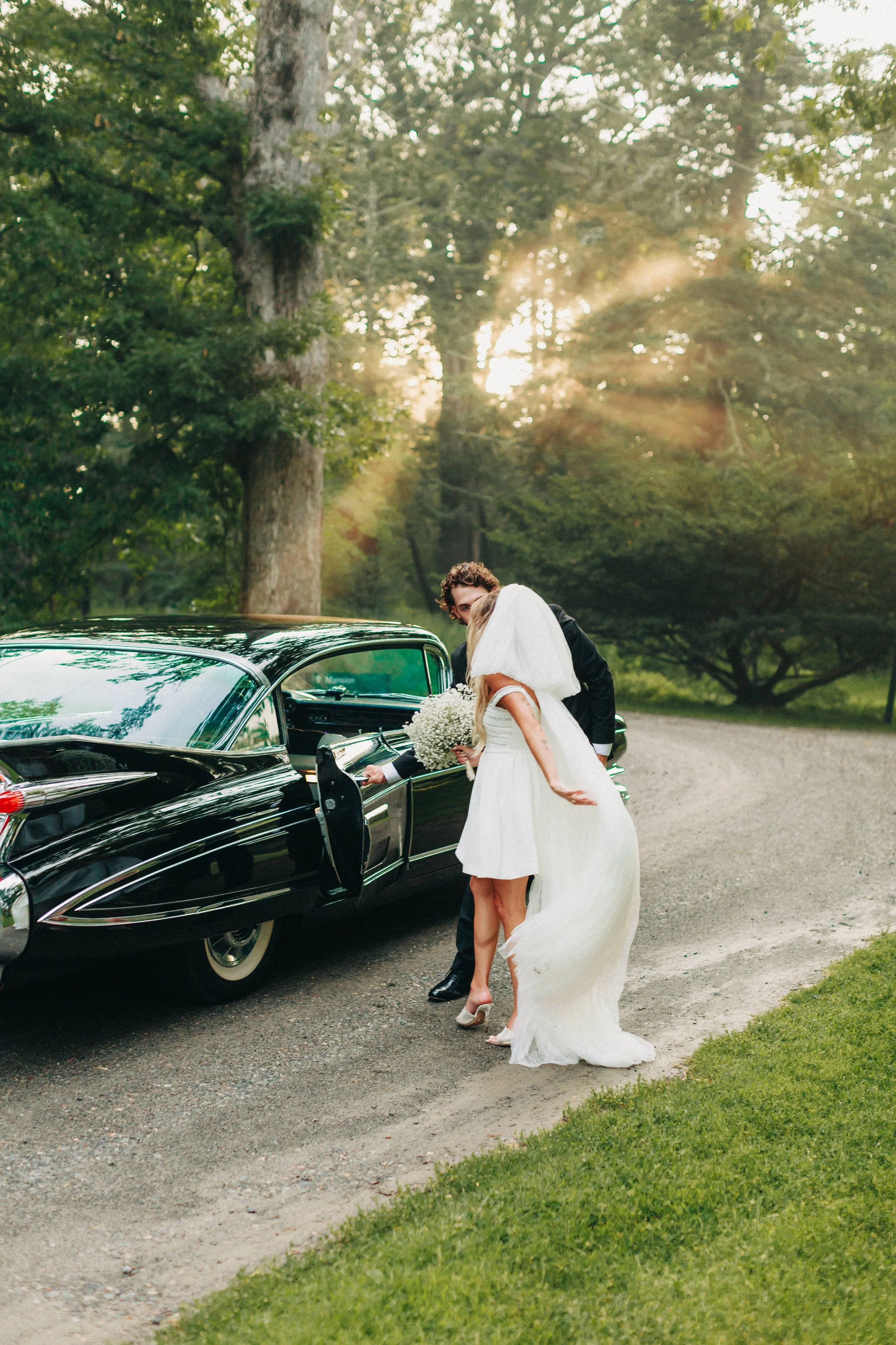 bride throws her beil behind her while groom opens the door of a black vintage cadillac for her to enter during their elopement in the hudson valley