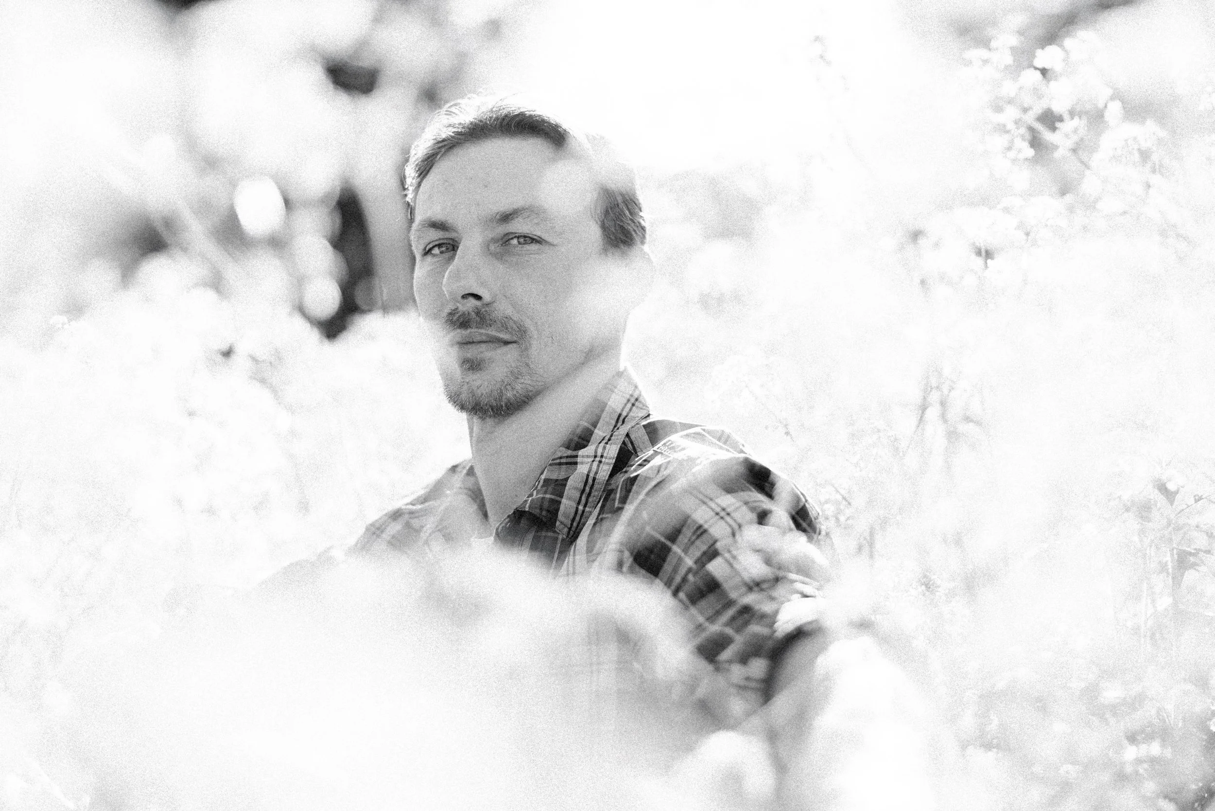 A black and white photo of a man with a short beard and slicked-back hair, wearing a plaid shirt, outdoors in a natural setting with trees and plants around him by Caitlinn Mahar-Daniels, hudson valley wedding photographer.