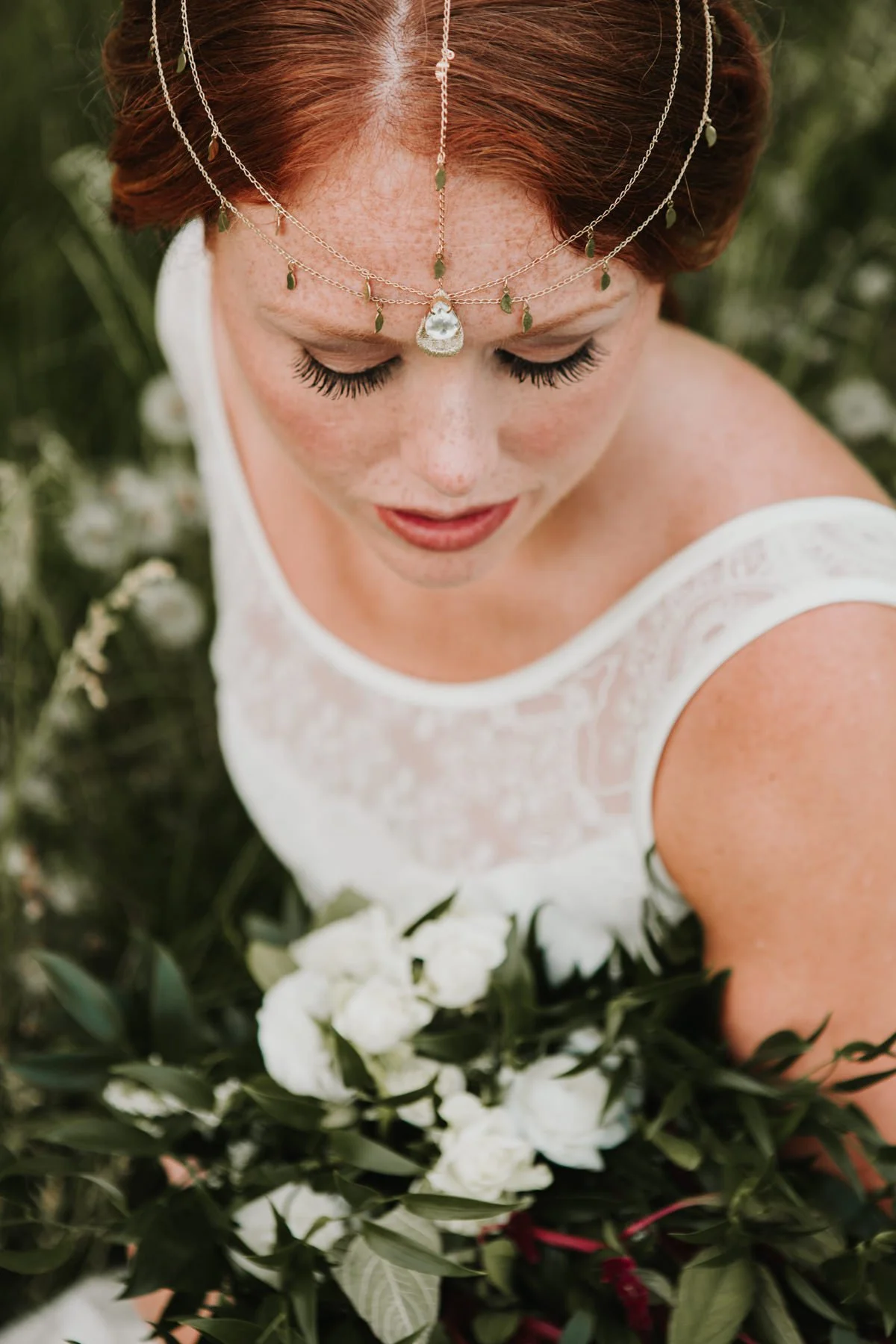 portrait of red haired bride wearing a beautiful headpiece while looking down sitting in the tall grass on her hudson valley wedding day