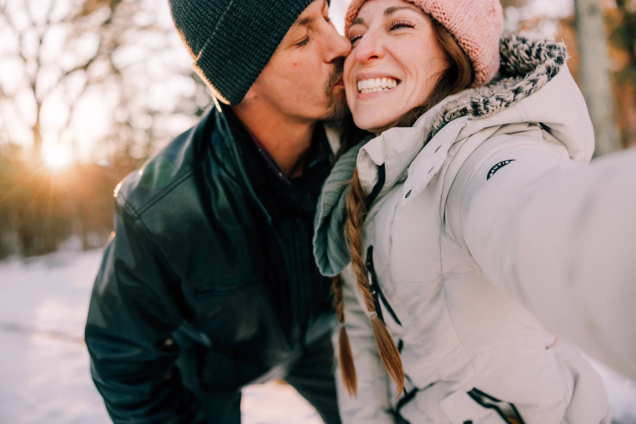 Caitlinn Mahar-Daniels, hudson valley wedding photographer, and her husband sharing a kiss outdoors in a snowy landscape during sunset, dressed in winter clothing at Franny Reese State Park.