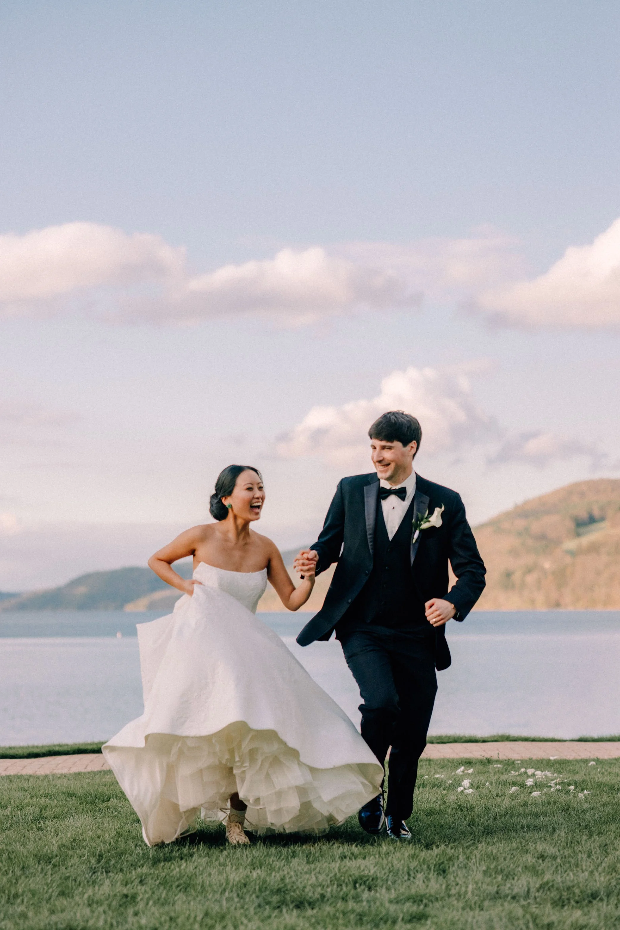 bride and groom holding hands while running and laughing on their wedding day with otsego lake behind them