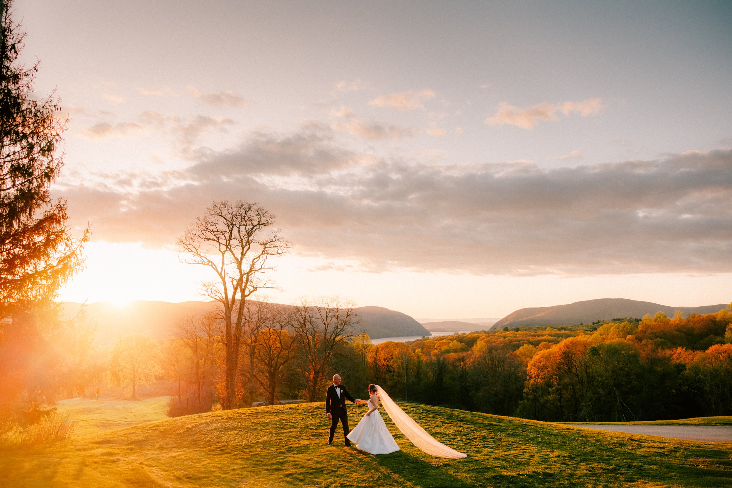bride and groom walking across hill at golden hour with the hudson river valley behind them on their wedding day in garrison new york