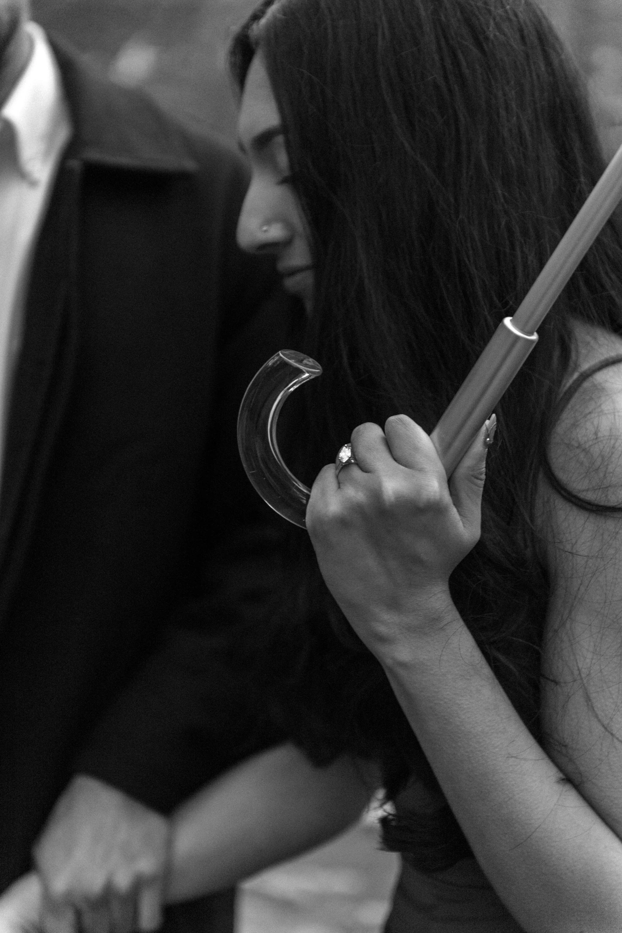 A woman with long dark hair holds a transparent umbrella, and a man nearby wears a dark jacket. The woman has a nose piercing and a ring on her finger. They were just engaged at Mohonk Mountain House.