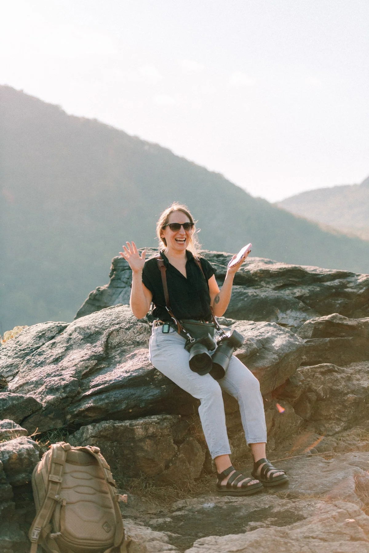 Caitlinn Mahar-Daniels, hudson valley wedding photographer, smiling, waving, holding a phone, with a camera hanging from her neck and surrounded by mountains at Little Stony Point waiting for her surprise proposal couple to arrive.