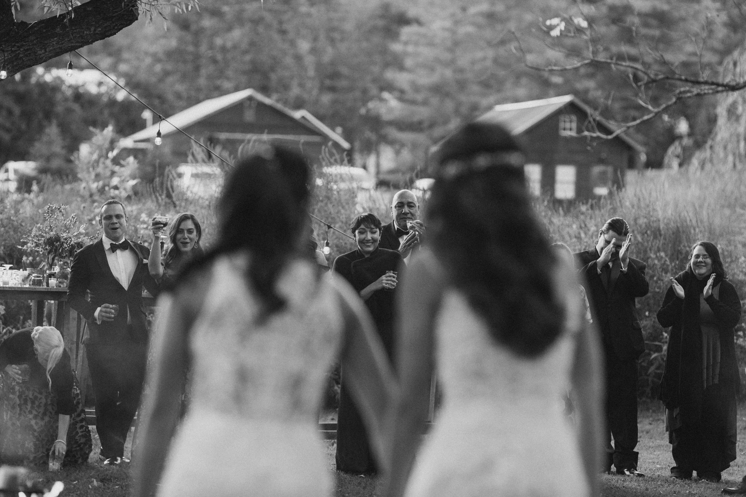 same sex couple hold hands after finishing their first dance at owls hoot barn while their intimate wedding guests cheer them on