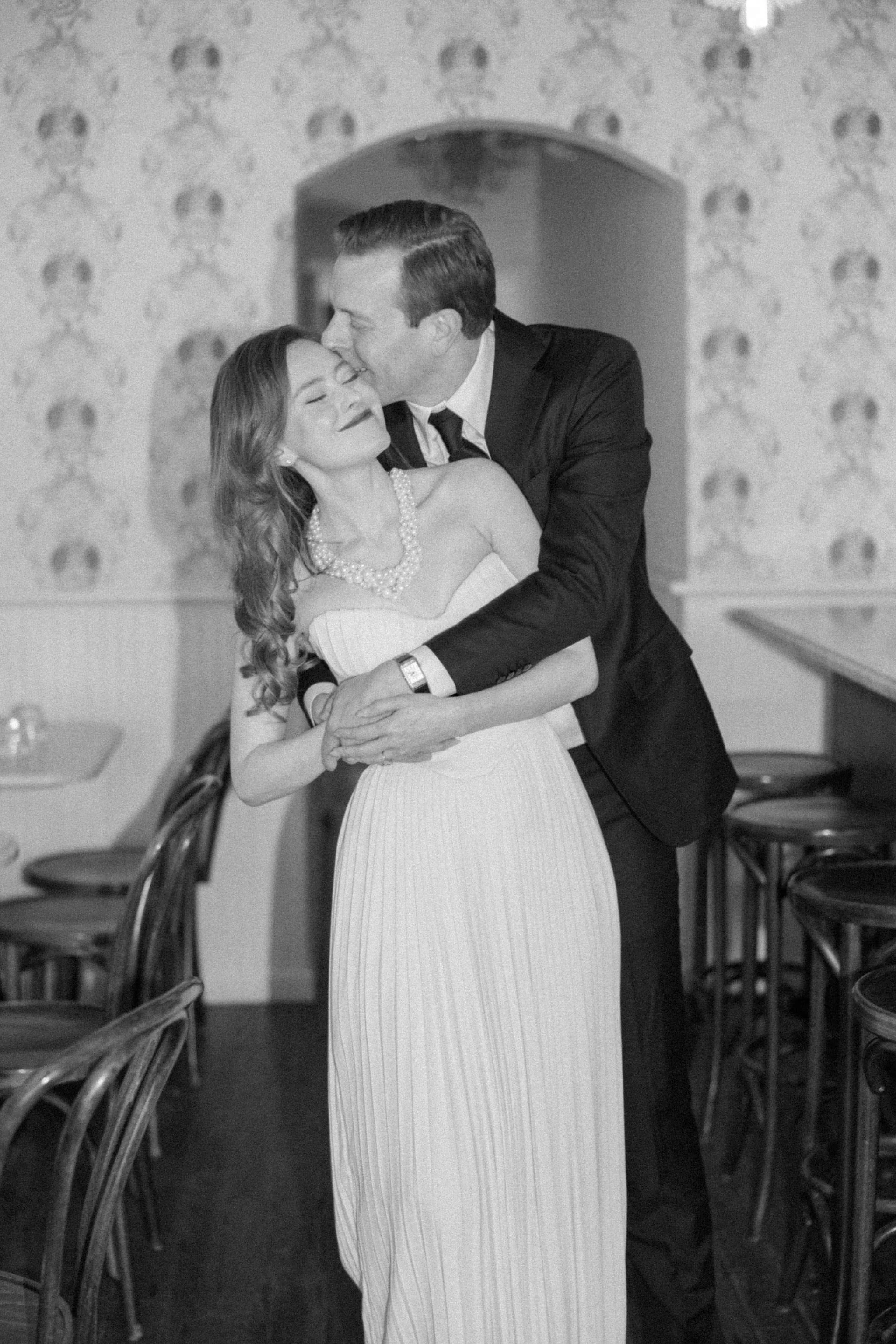film photograph of groom hugging his future bride from behind she is wearing a white strapless dress and a pearl necklace in brunette wine bar
